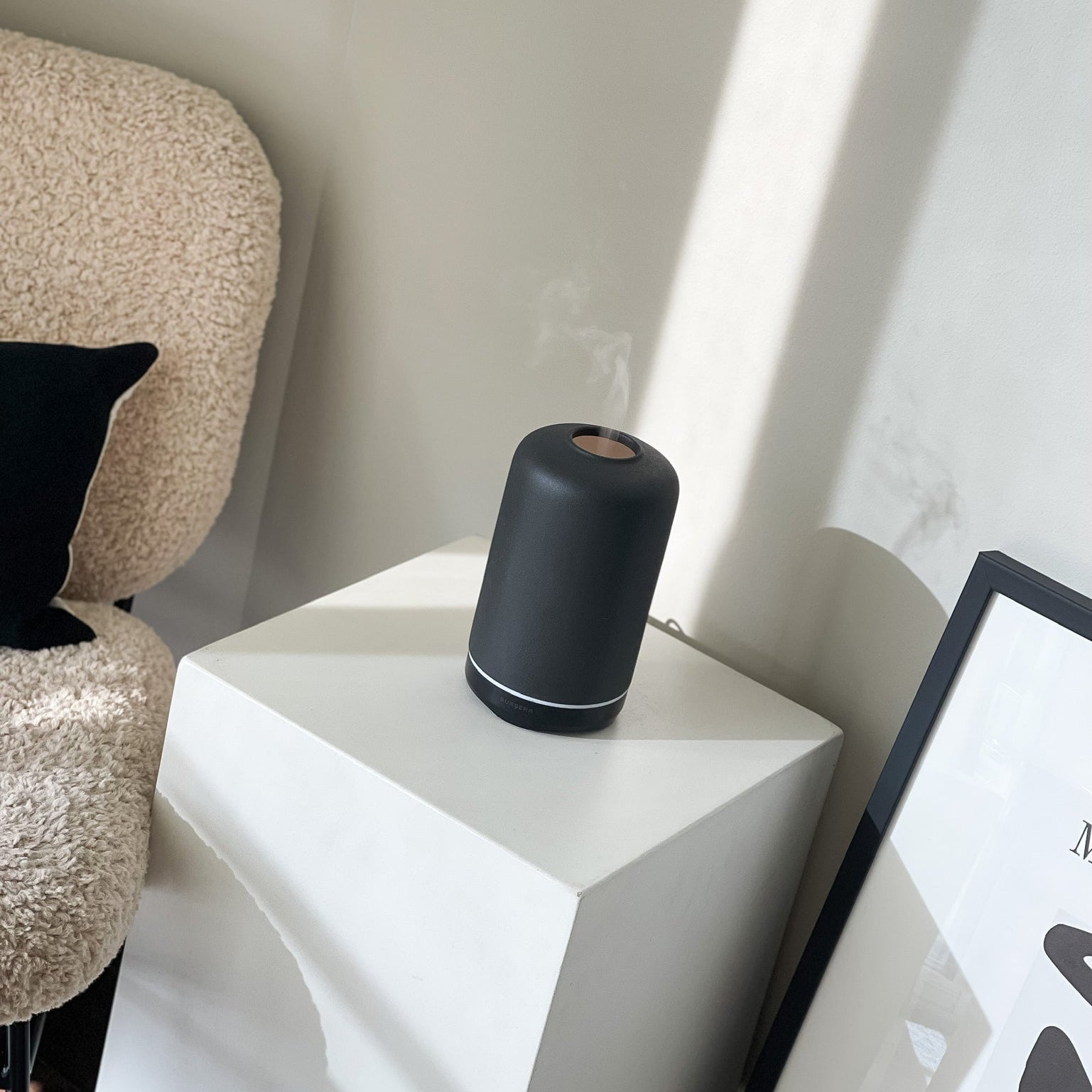 Black cylindrical humidifier on a white geometric side table with a textured chair and framed picture in the background.