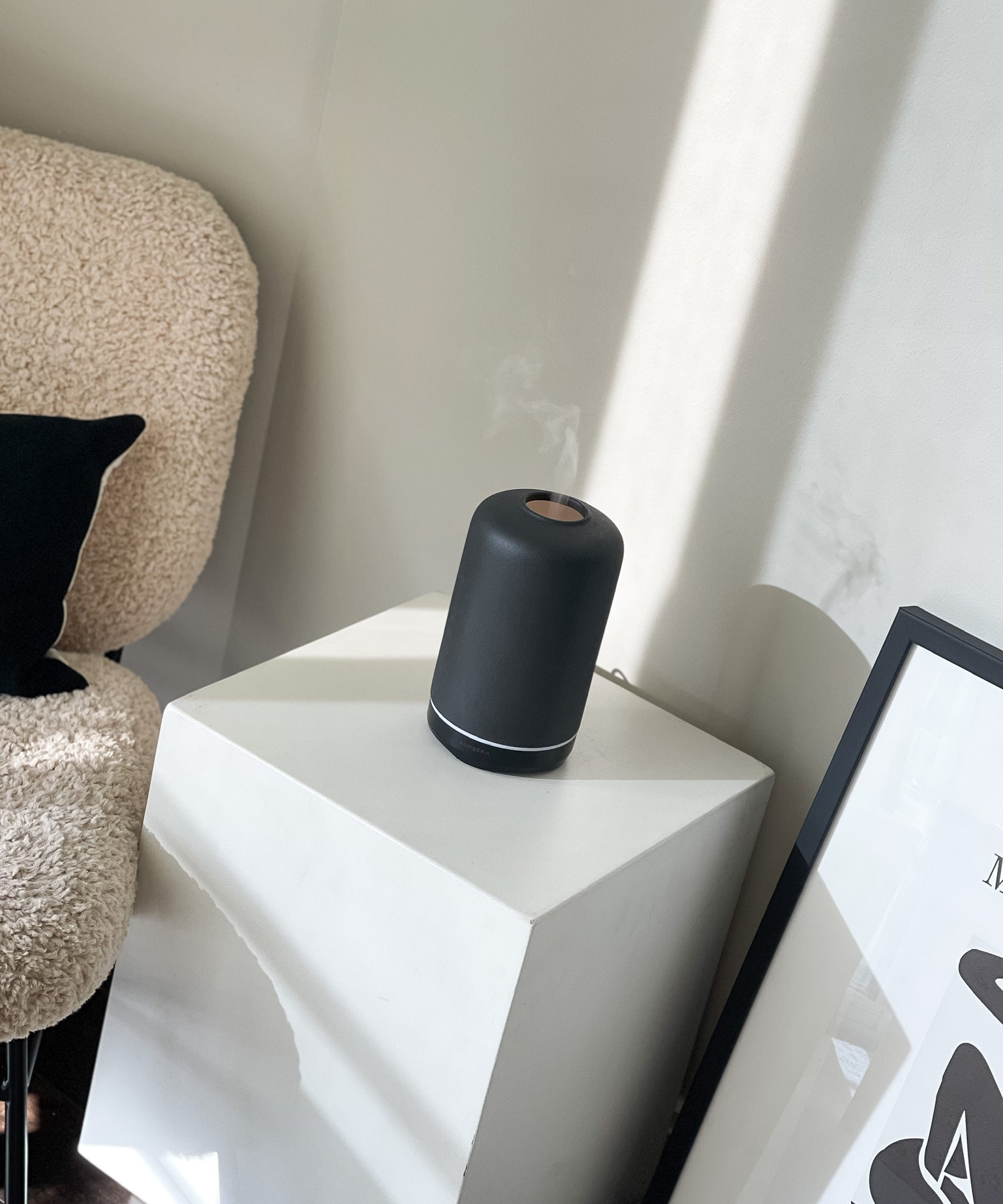 Black cylindrical humidifier on a white geometric side table with a textured chair and framed picture in the background.