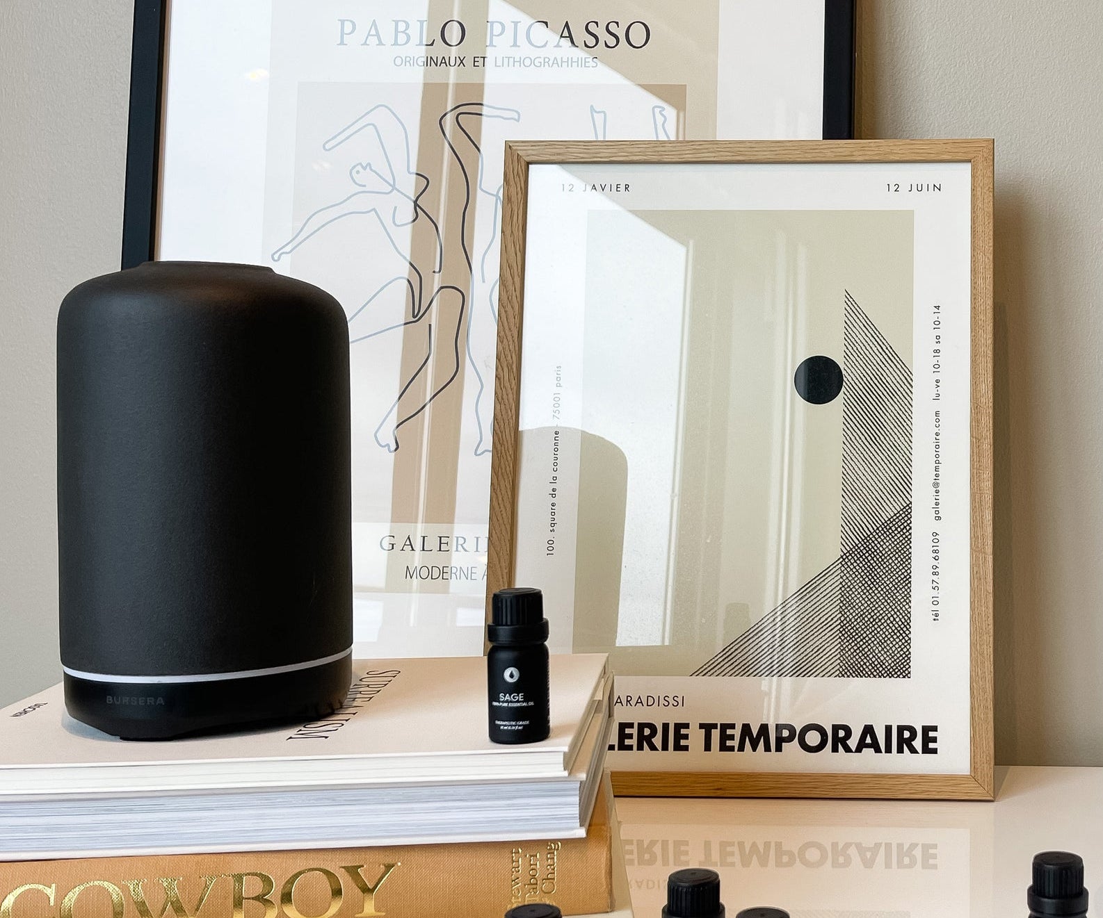Black cylindrical product, framed art prints, and small black bottles on a surface.