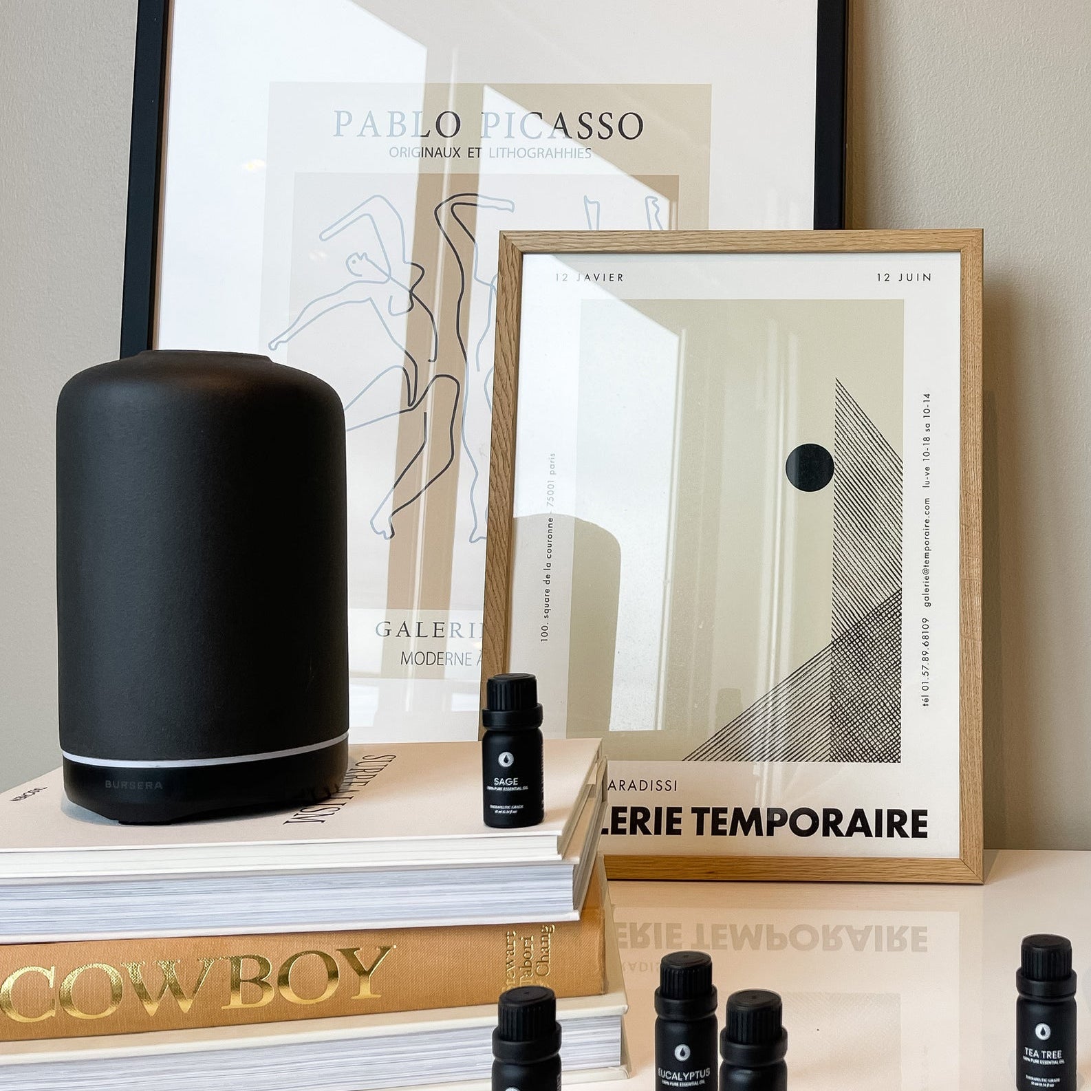 Black cylindrical product, framed art prints, and small black bottles on a surface.