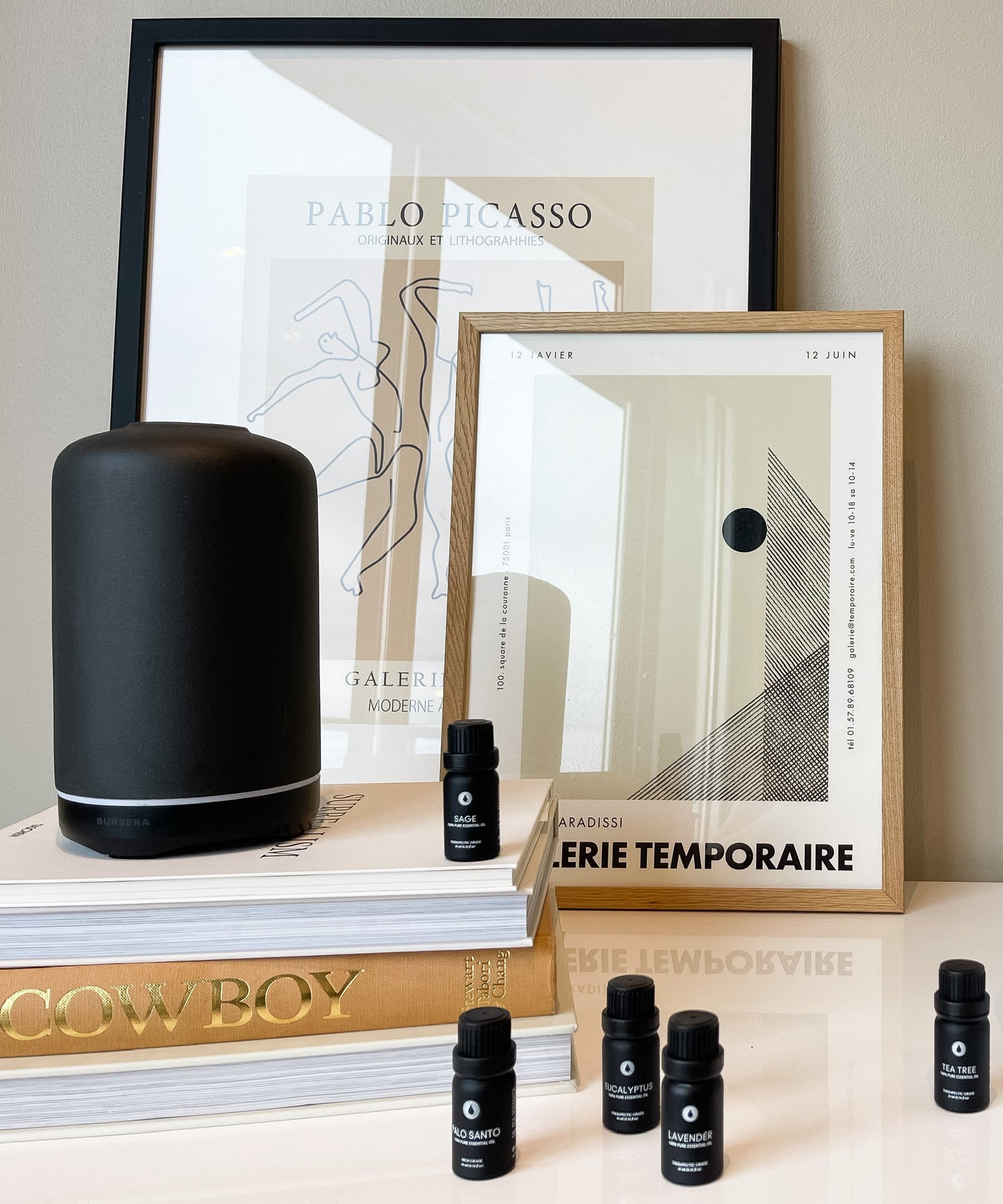 Black cylindrical product, framed art prints, and small black bottles on a surface.
