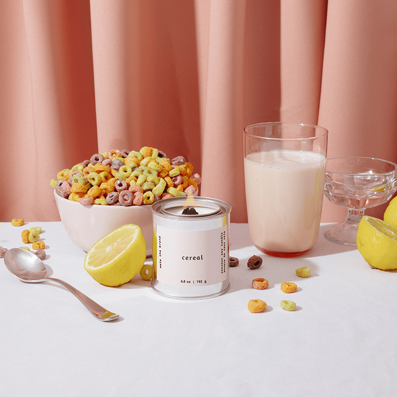 Cereal bowl with colorful cereal, a glass of milk, and a container labeled 'cereal' on a white surface with pink curtains in the background.