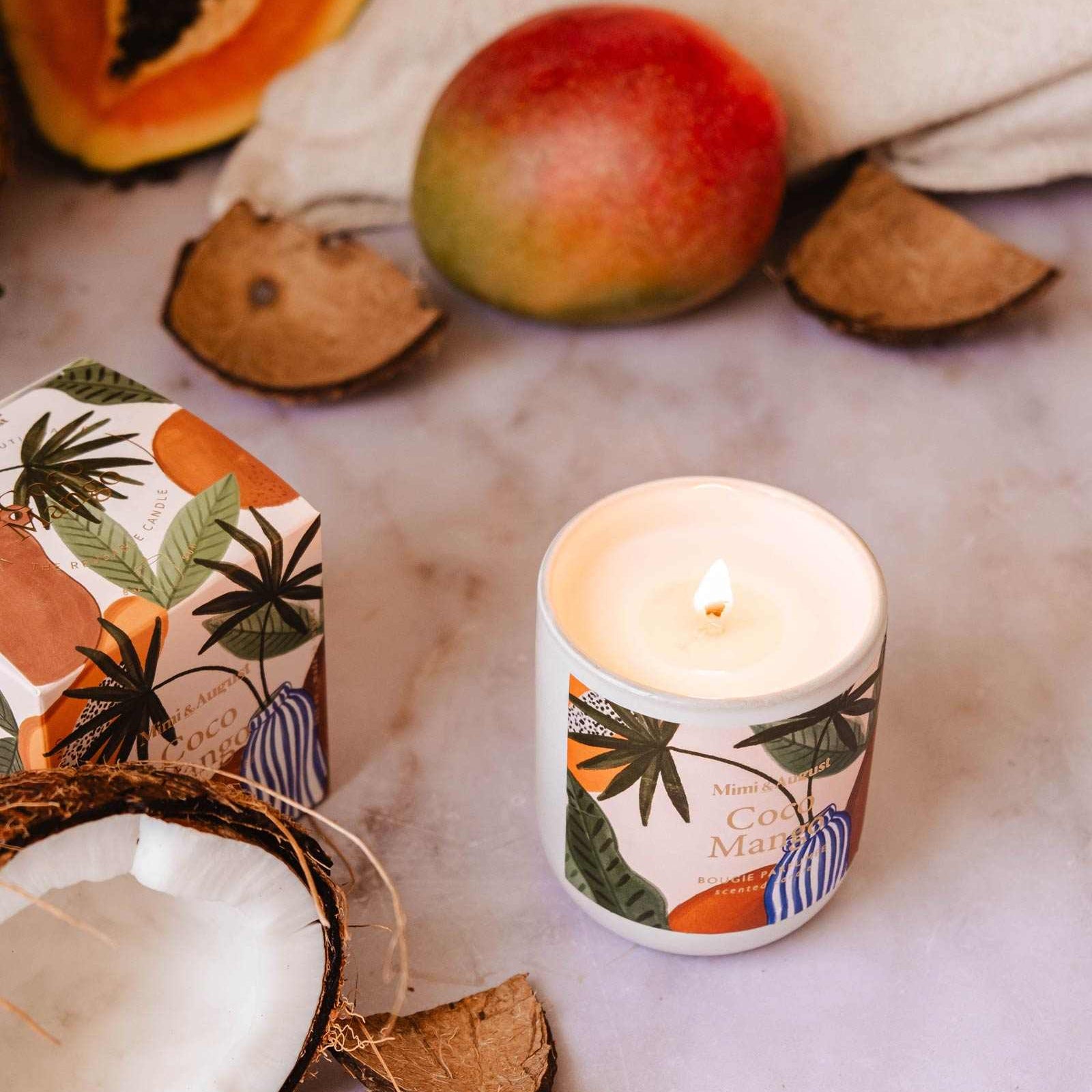 Candle with coconut and mango design surrounded by coconuts and a mango on a light background