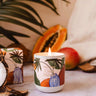 Candle with tropical design surrounded by fruits and leaves on a textured surface