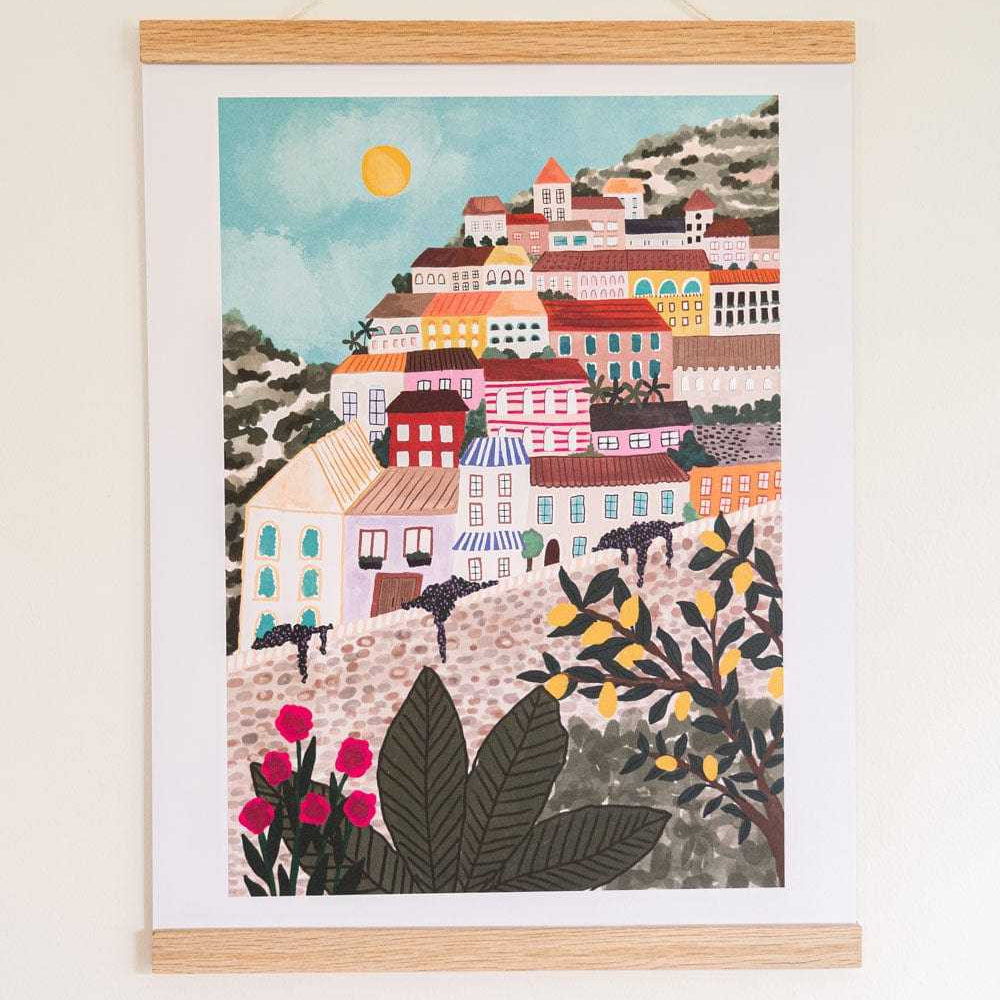 A framed art print depicting a colorful hillside village scene with buildings, plants, and a sun, hung on a light wall.