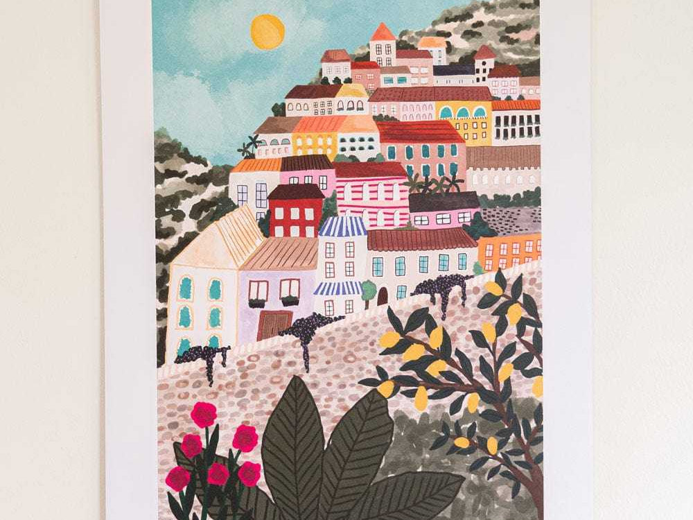 A framed art print depicting a colorful hillside village scene with buildings, plants, and a sun, hung on a light wall.