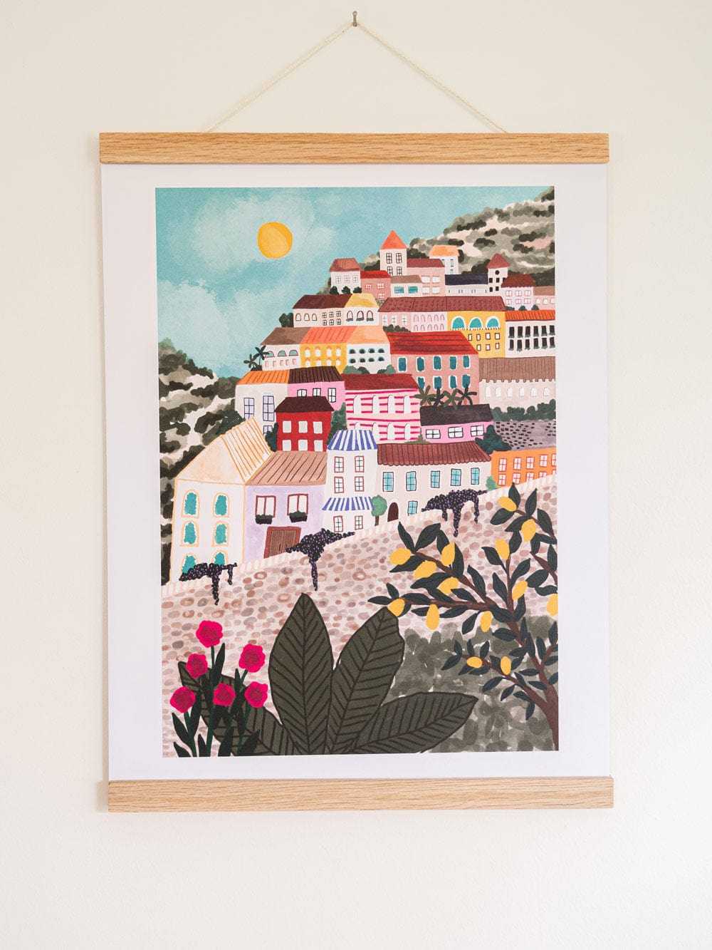 A framed art print depicting a colorful hillside village scene with buildings, plants, and a sun, hung on a light wall.