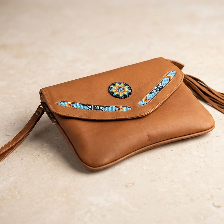 OldTribes A tan leather crossbody bag with a flap closure, decorated with a colorful beaded circular medallion and matching beaded arrow details on the flap, resting on a light beige textured surface.