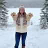 A woman standing in a snowy outdoor setting with trees in the background, wearing a cream knit sweater, a multicolor knit scarf, a pink beanie, and oversized beige mittens shaped like animal paws.