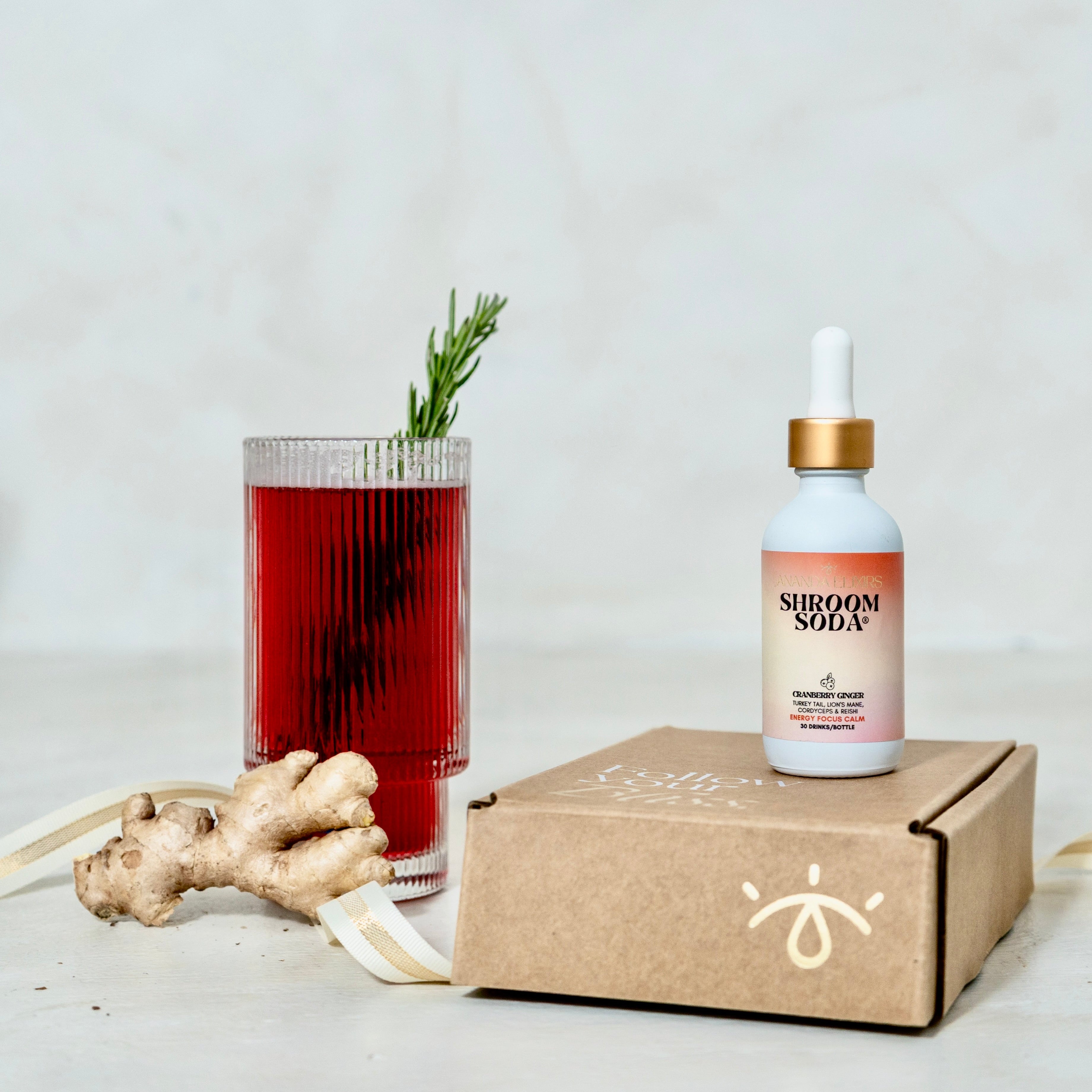 Ananda Elixirs Shroom Soda® – Cranberry Ginger – Mocktail in ribbed glass with Ananda Elixirs gift box and fresh ginger