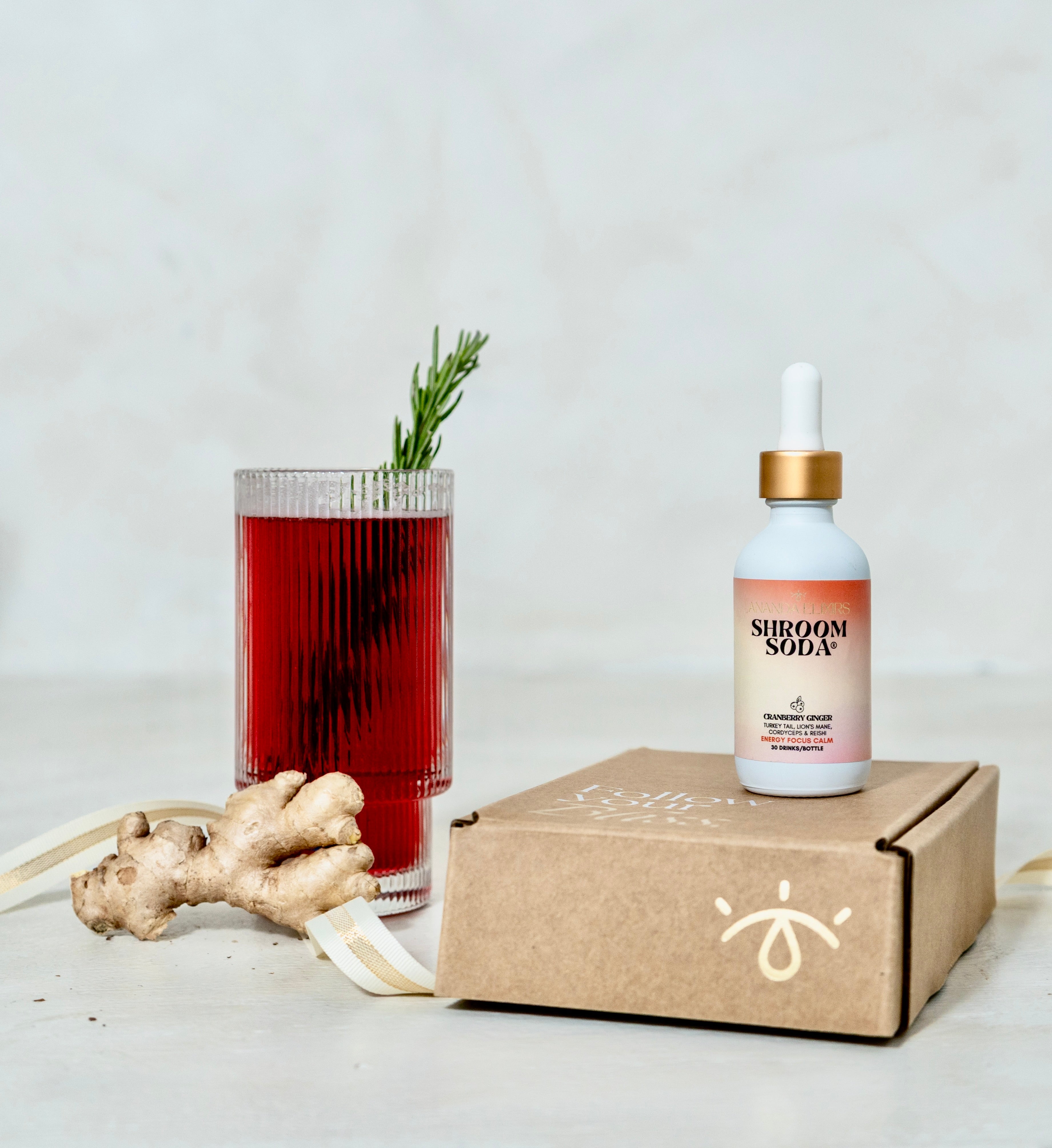 Ananda Elixirs Shroom Soda® – Cranberry Ginger – Mocktail in ribbed glass with Ananda Elixirs gift box and fresh ginger