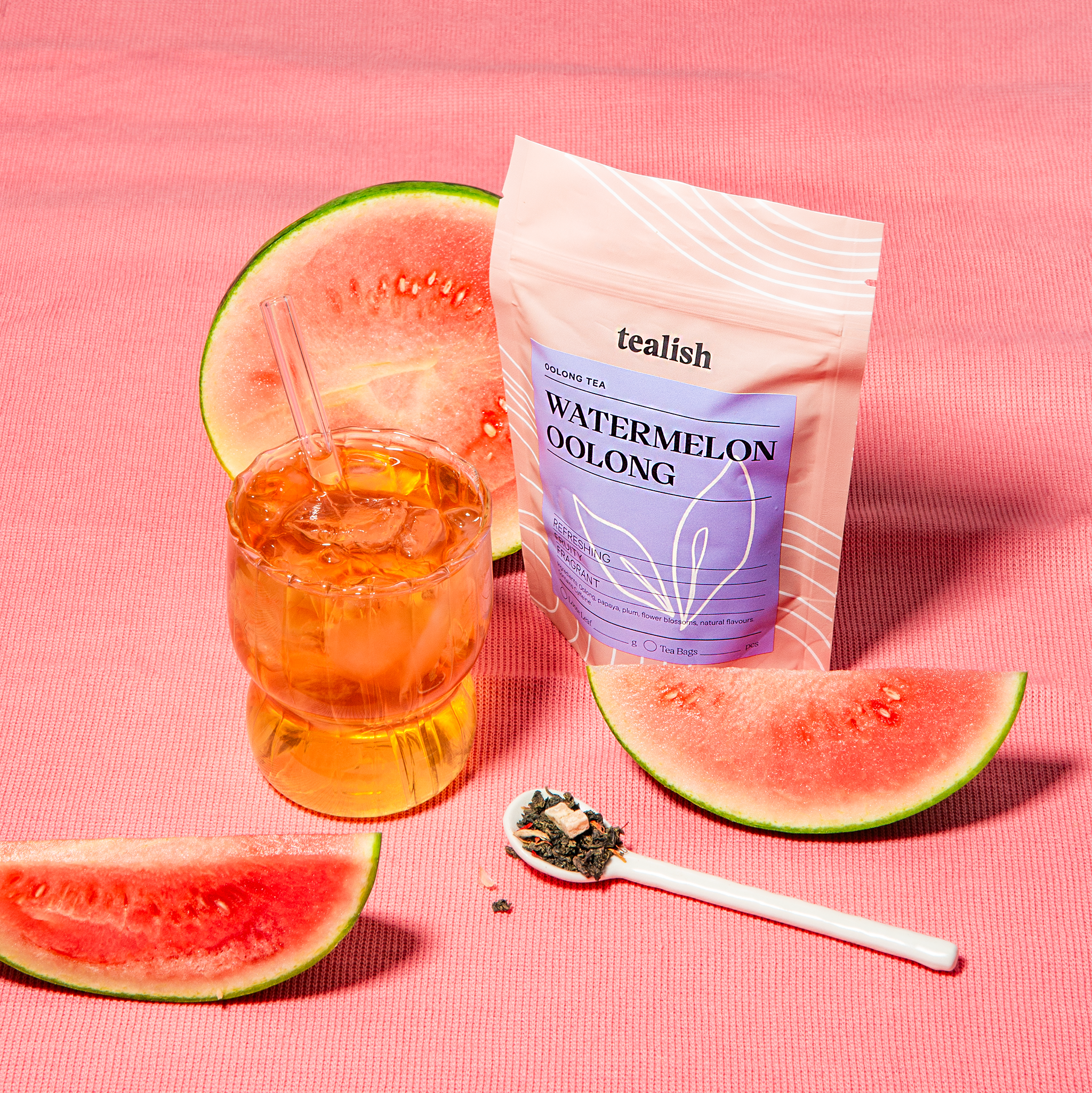 Tealish Watermelon Oolong tea package with a glass of iced tea, watermelon slices, and a spoonful of tea leaves on a pink background.