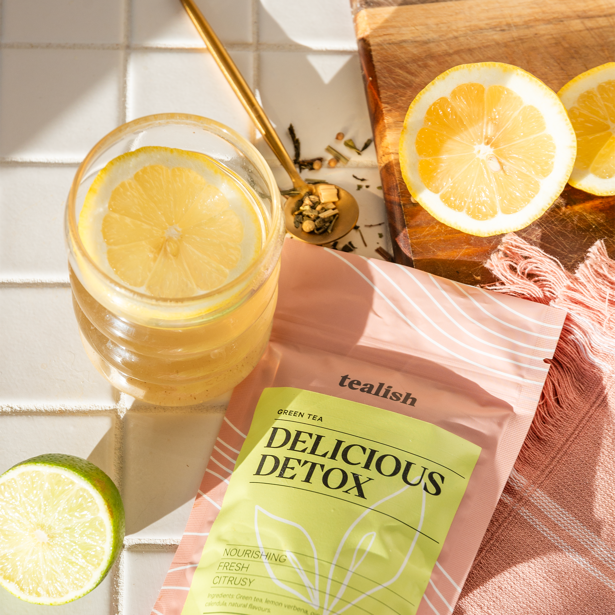Tealish Delicious Detox tea package with a glass of lemon water and lemon slices on a wooden cutting board.