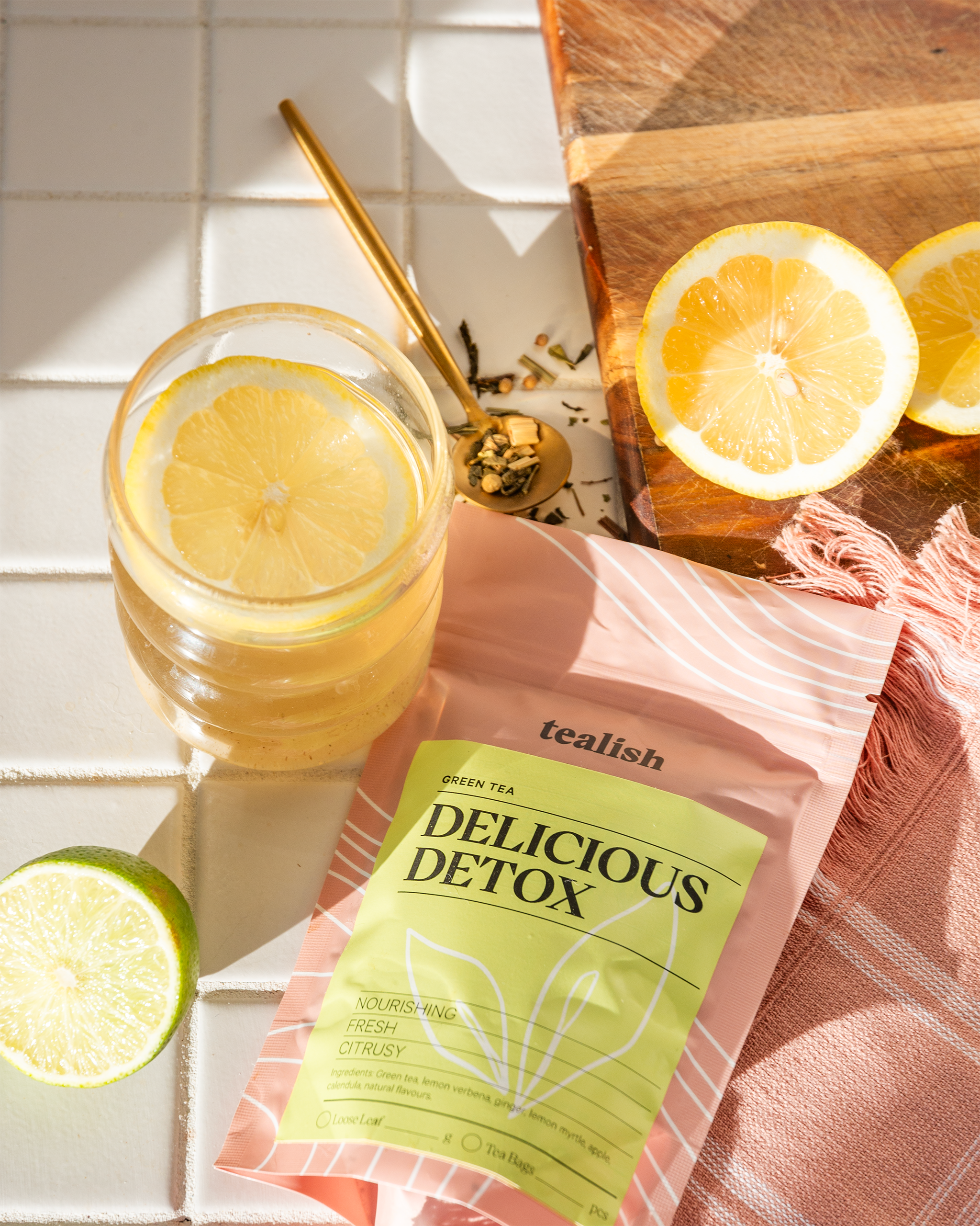 Tealish Delicious Detox tea package with a glass of lemon water and lemon slices on a wooden cutting board.