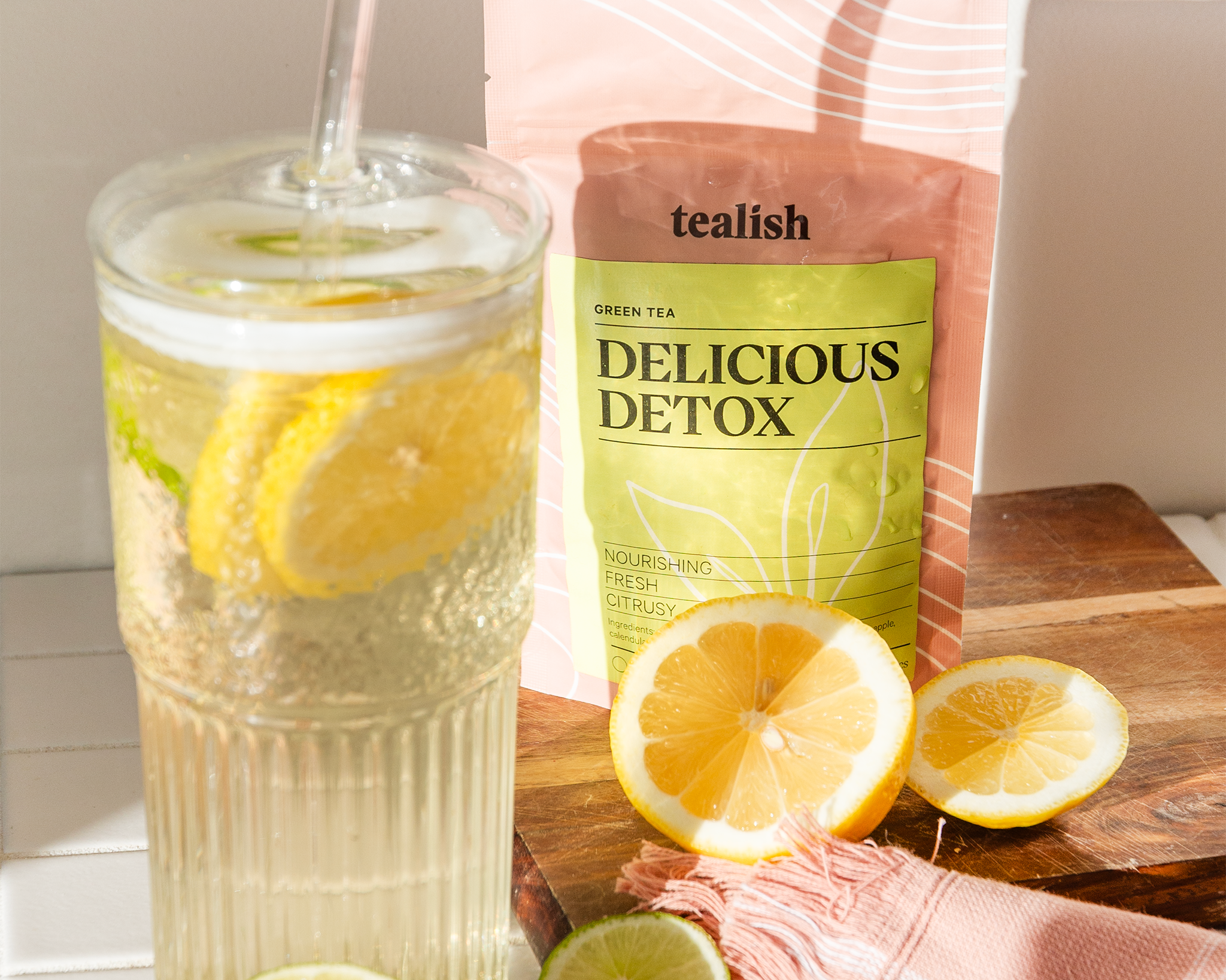 Glass of lemon water with a tealish brand 'Delicious Detox' package on a wooden surface.
