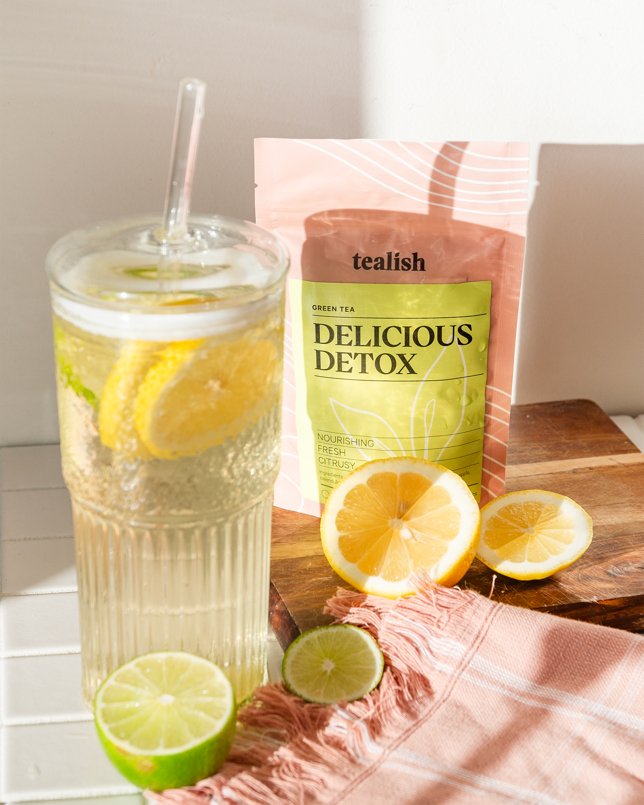 Glass of lemon water with a tealish brand 'Delicious Detox' package on a wooden surface.