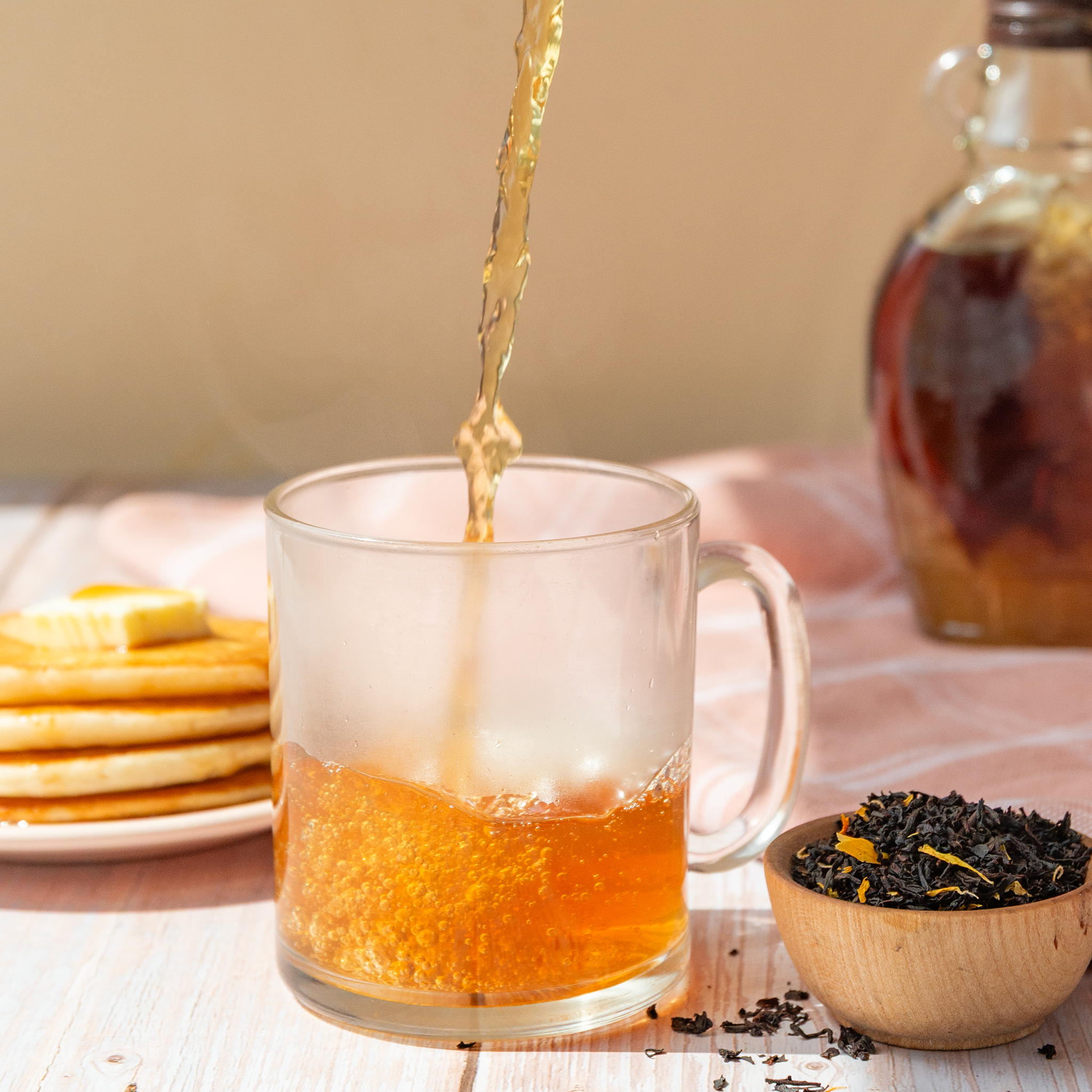 Tea being poured into a glass mug with pancakes and a jar of syrup in the background.