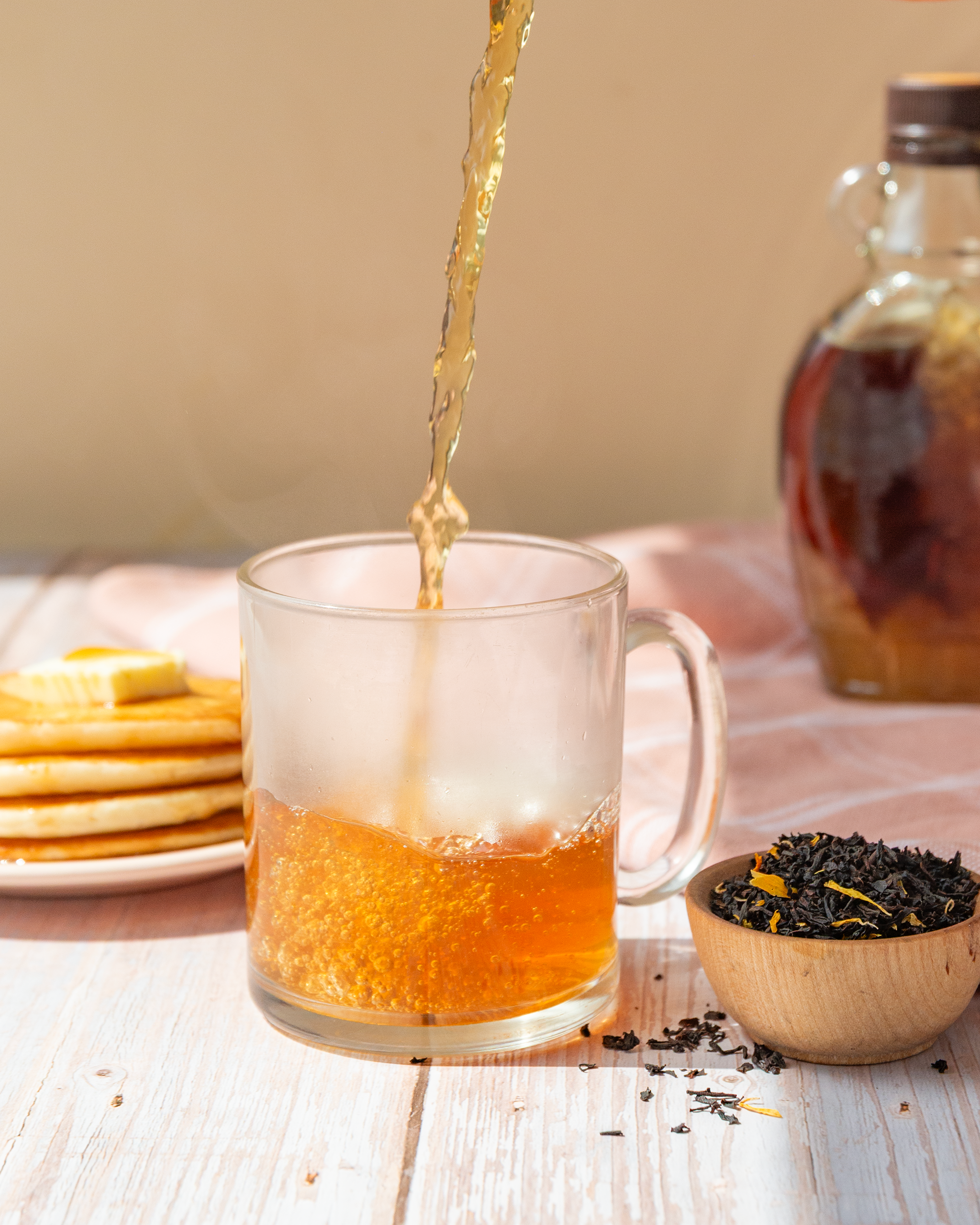 Tea being poured into a glass mug with pancakes and a jar of syrup in the background.