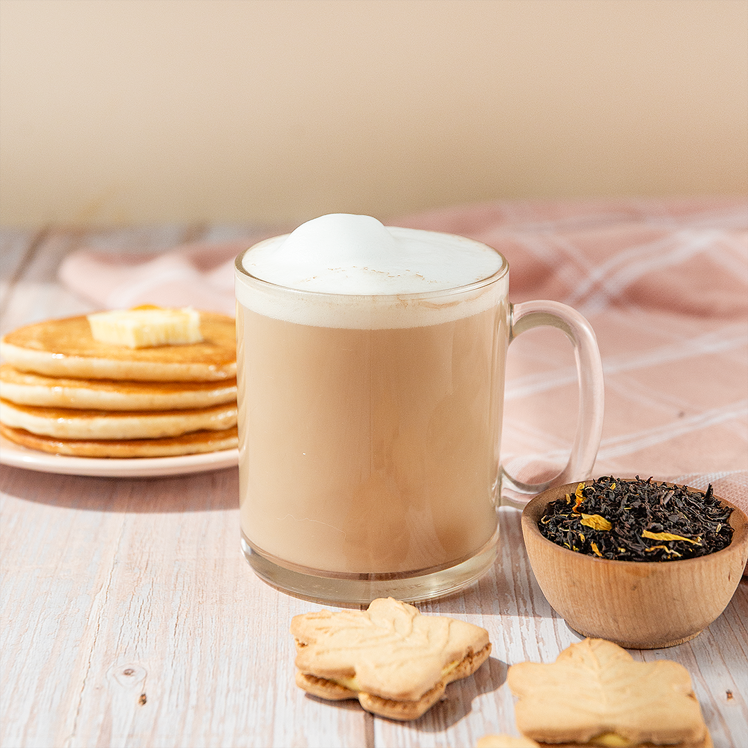 Glass mug with creamy beverage, cookies, and a bowl of tea leaves on a wooden surface.