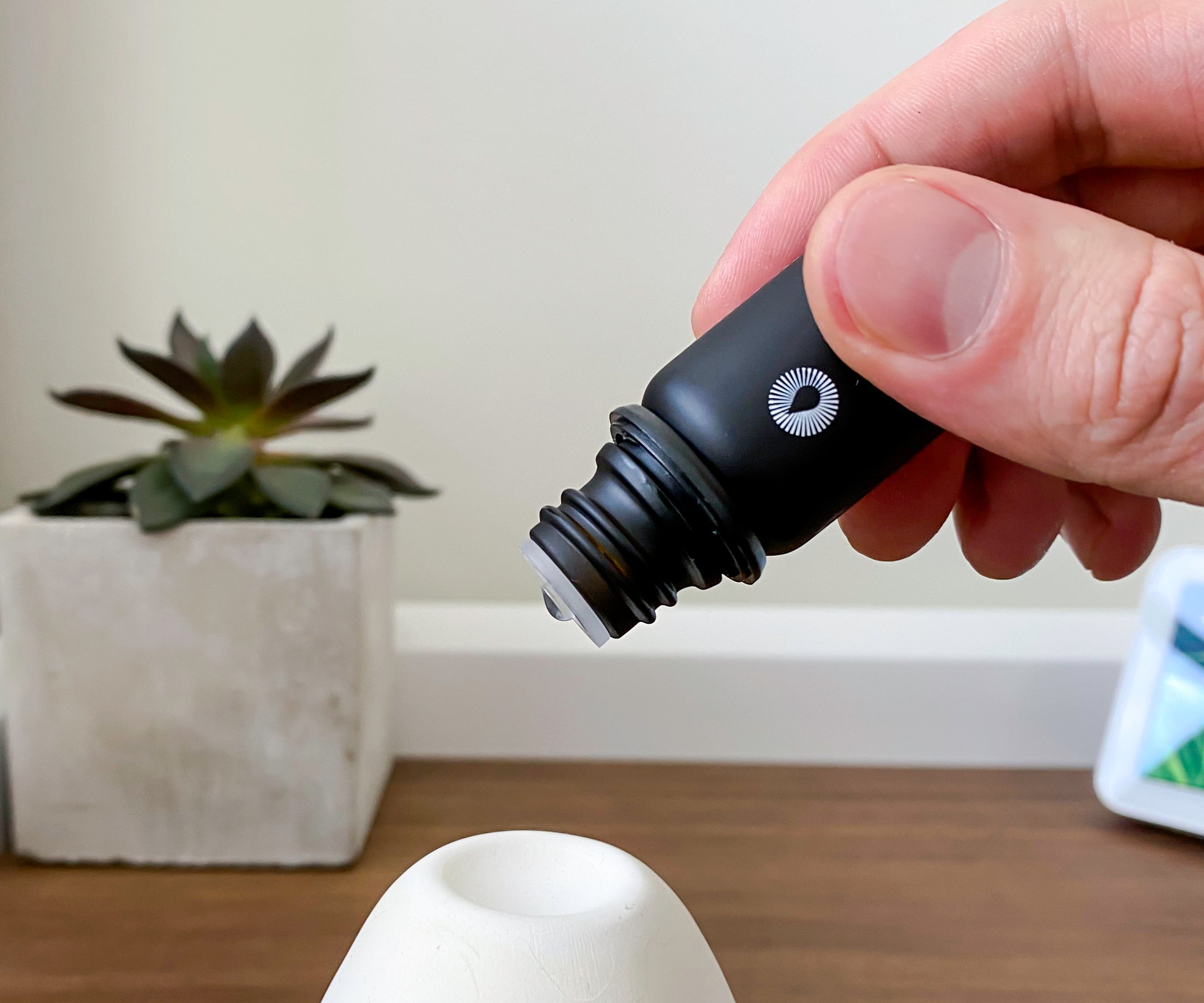 Hand holding a black essential oil dropper near a white ceramic object on a wooden surface with a plant in the background.