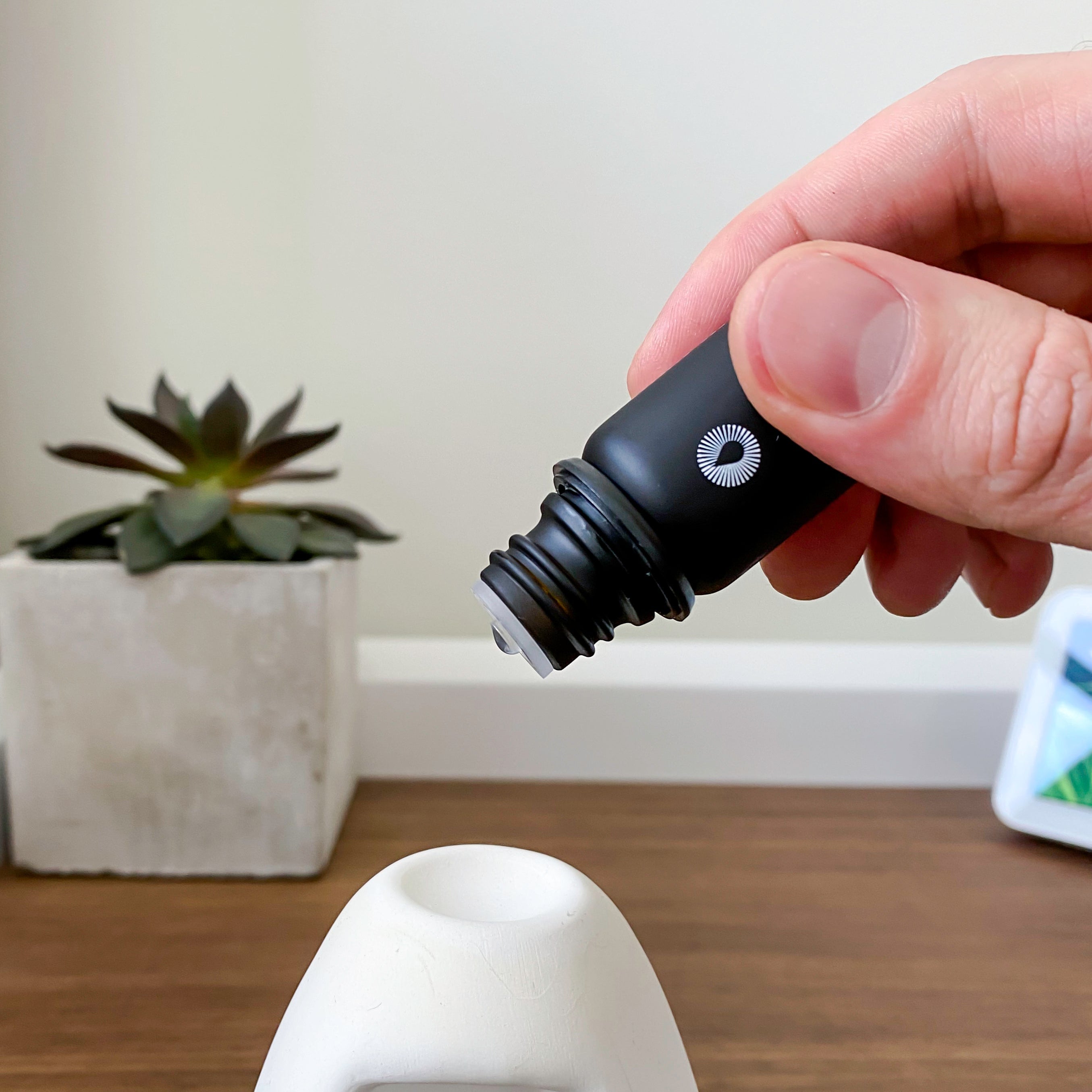 Hand holding a black essential oil dropper near a white ceramic object on a wooden surface with a plant in the background.