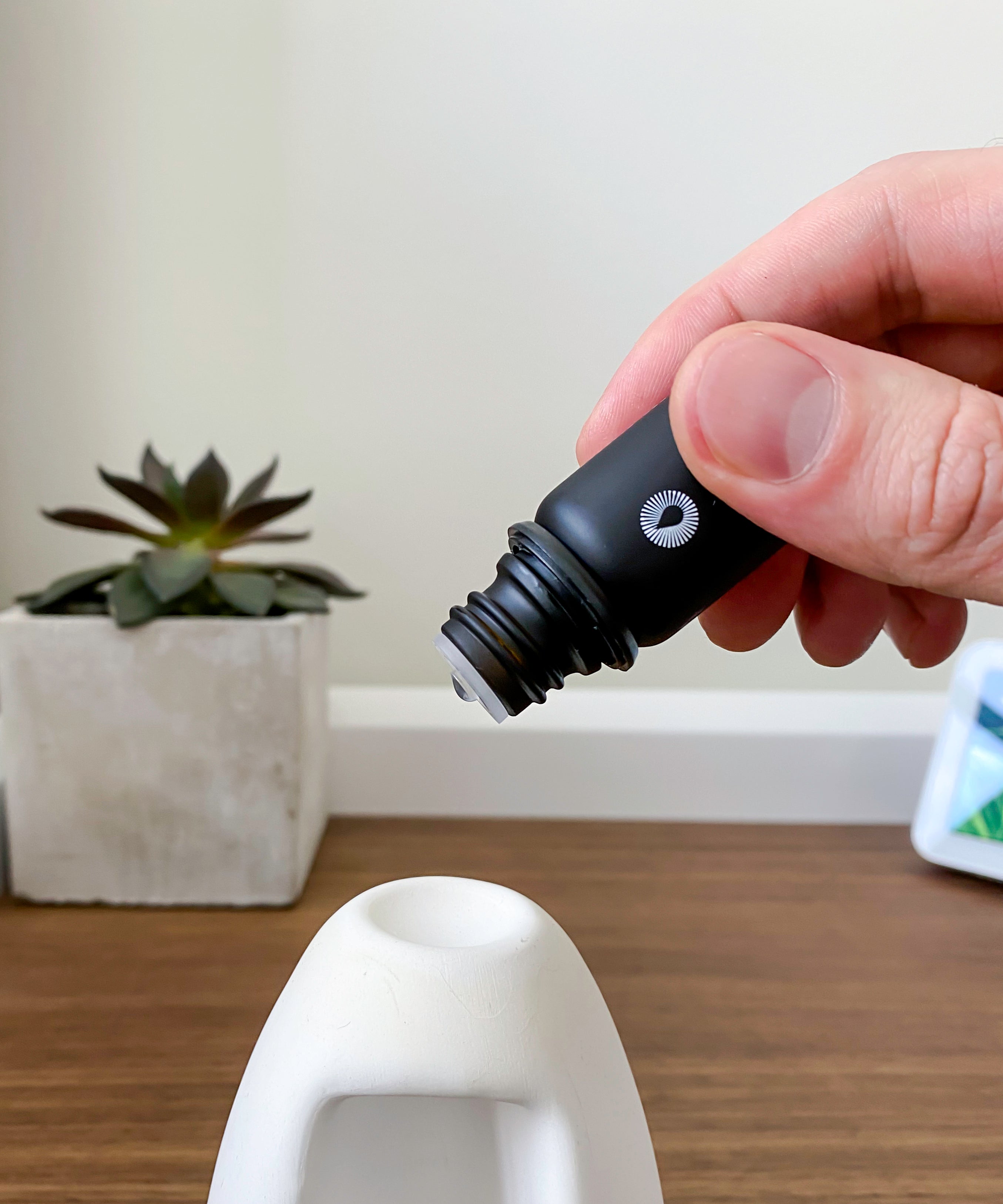 Hand holding a black essential oil dropper near a white ceramic object on a wooden surface with a plant in the background.