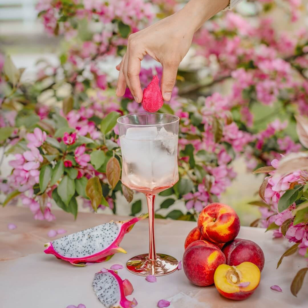 Hand dipping a pink flower into a glass of water with dragon fruit and peaches on a floral background