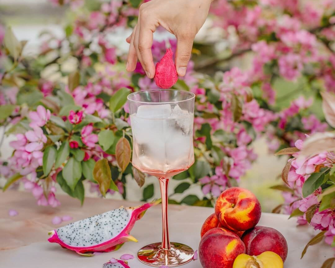 Hand dipping a pink flower into a glass of water with dragon fruit and peaches on a floral background
