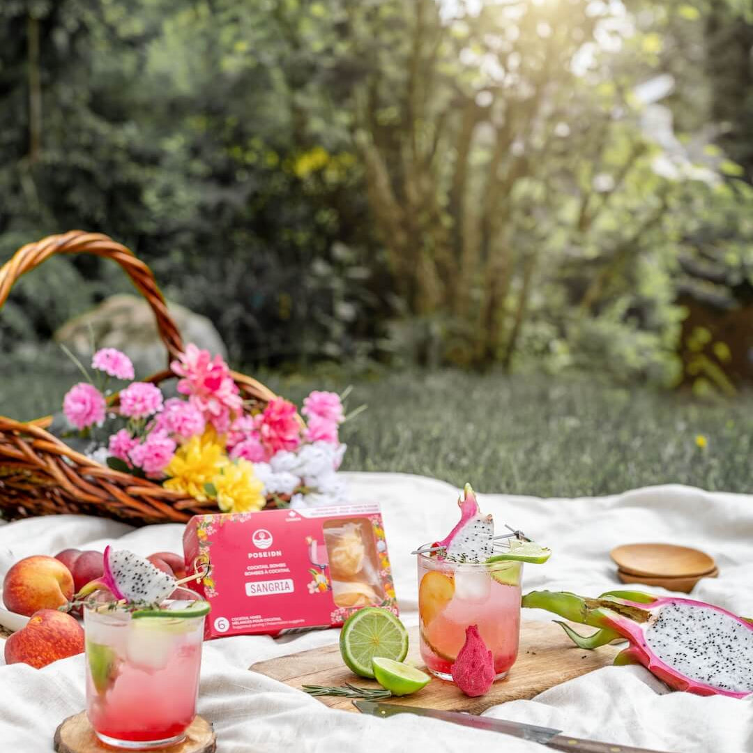 Outdoor picnic setup with drinks, fruits, and a box on a white blanket.
