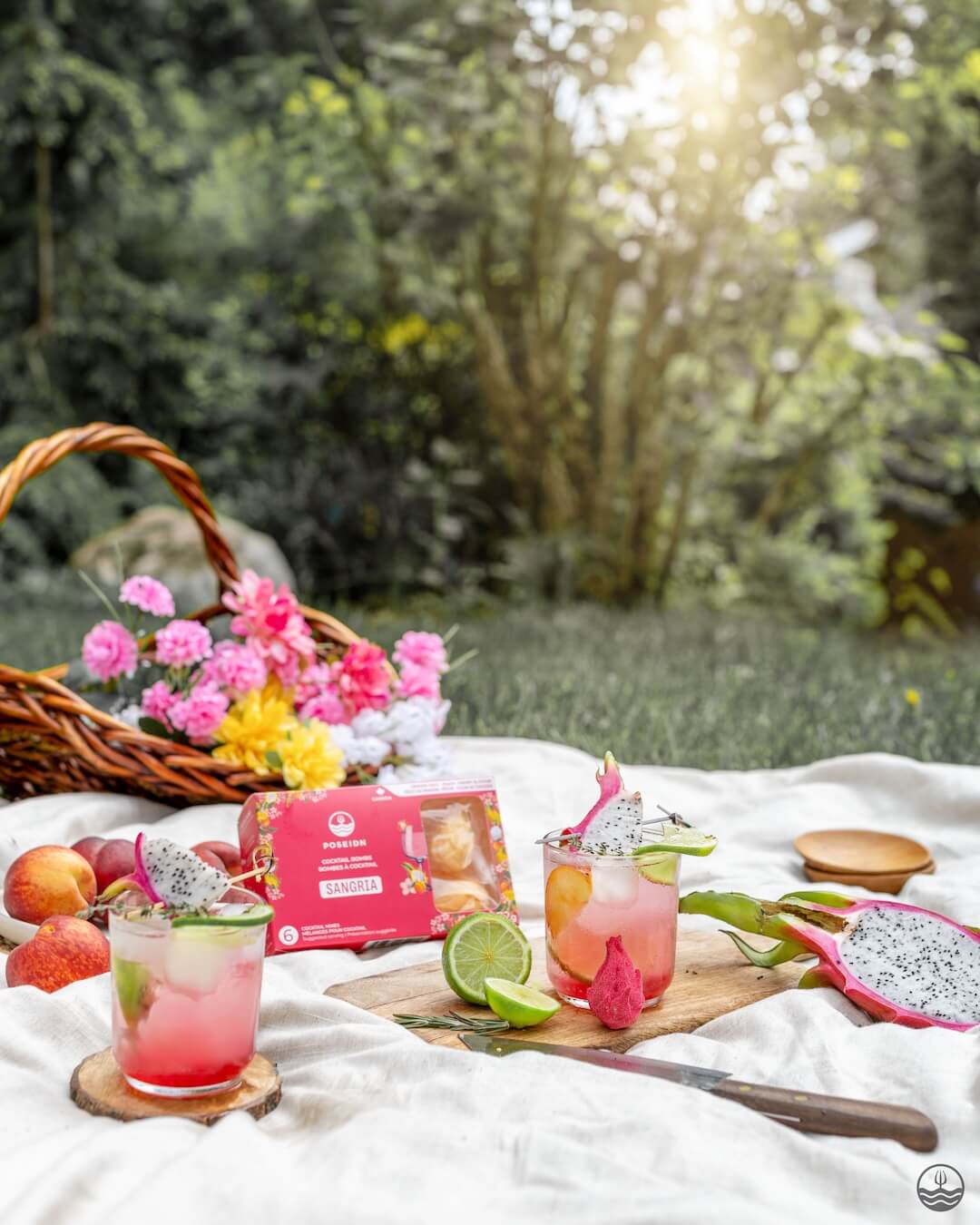 Outdoor picnic setup with drinks, fruits, and a box on a white blanket.