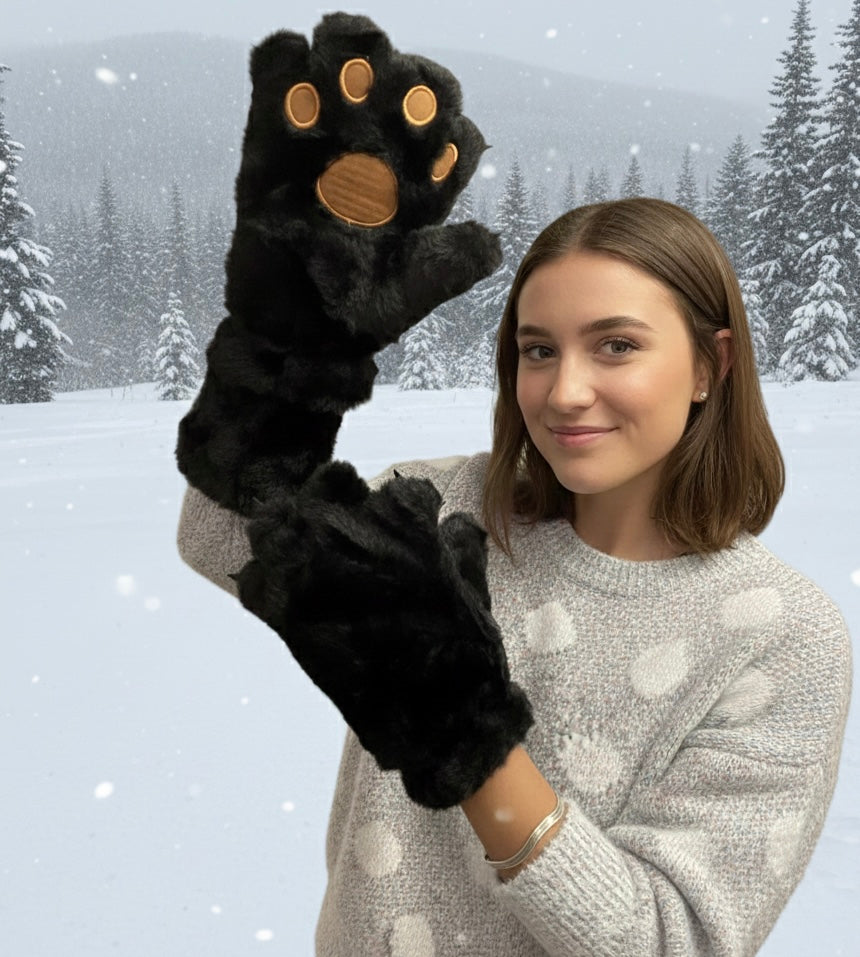 OldTribes Black Soft Plush Bear Paw Gloves – Cozy Winter Gloves A woman wearing a light gray knit sweater holds up a black plush winter glove shaped like a cat paw, with tan paw pads and claws, against a snowy forest background.