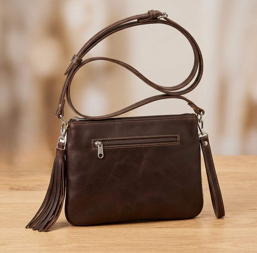 A dark brown leather crossbody handbag with a matching tassel and adjustable shoulder strap, resting on a light wooden surface with a warm, blurred background.
