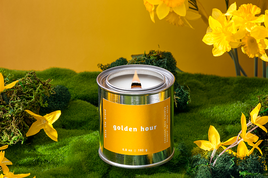 Candle labeled 'Golden Hour' surrounded by yellow flowers on a green surface with a yellow background