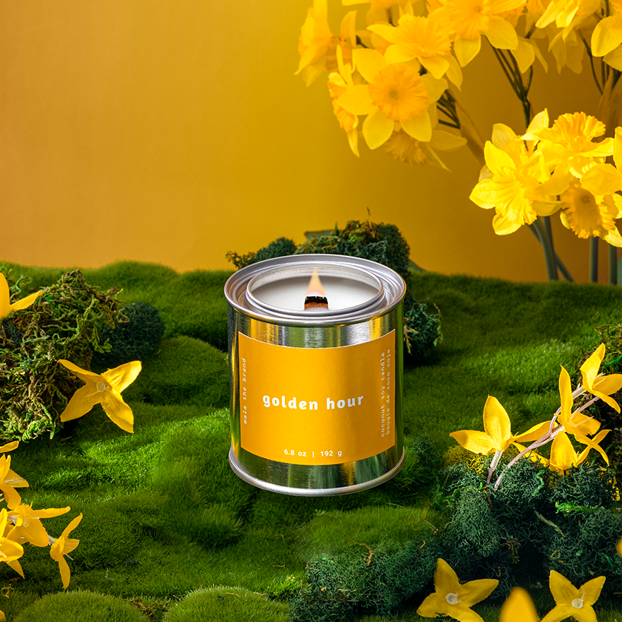 Candle labeled 'Golden Hour' surrounded by yellow flowers on a green surface with a yellow background