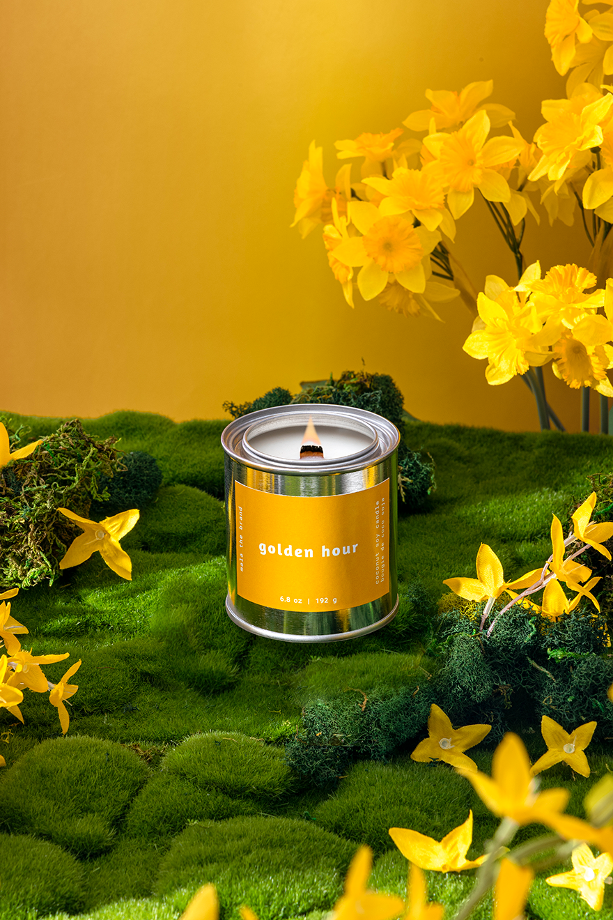 Candle labeled 'Golden Hour' surrounded by yellow flowers on a green surface with a yellow background