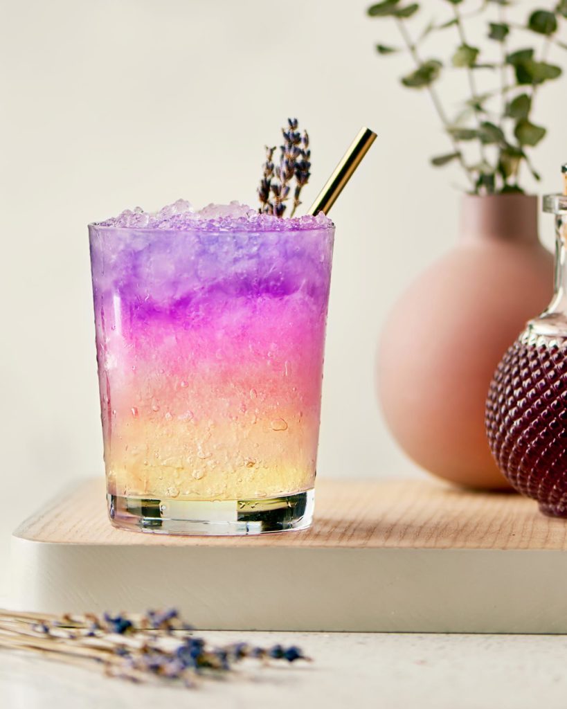 Colorful cocktail with ice and lavender garnish on a wooden surface with a light background The Sobr Market