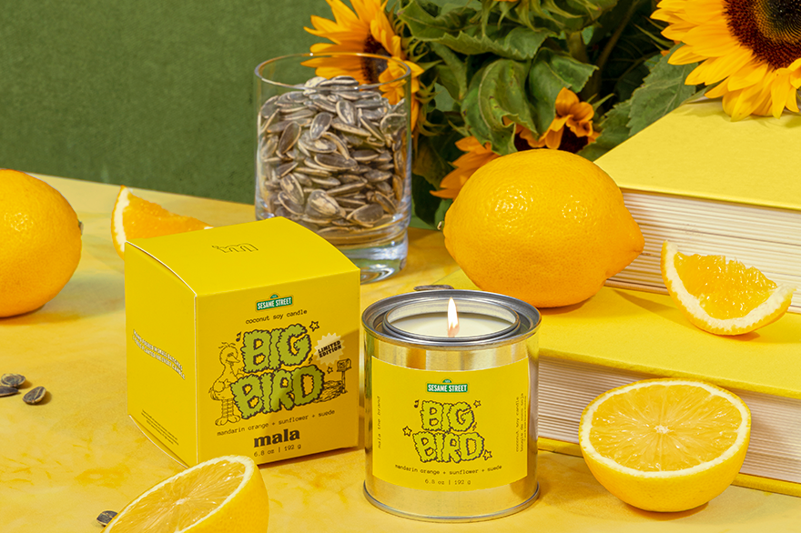 Yellow candle with sunflower seeds packaging on a table with sunflowers and oranges.