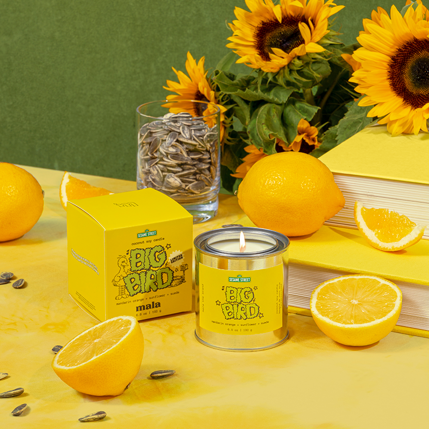 Yellow candle with sunflower seeds packaging on a table with sunflowers and oranges.