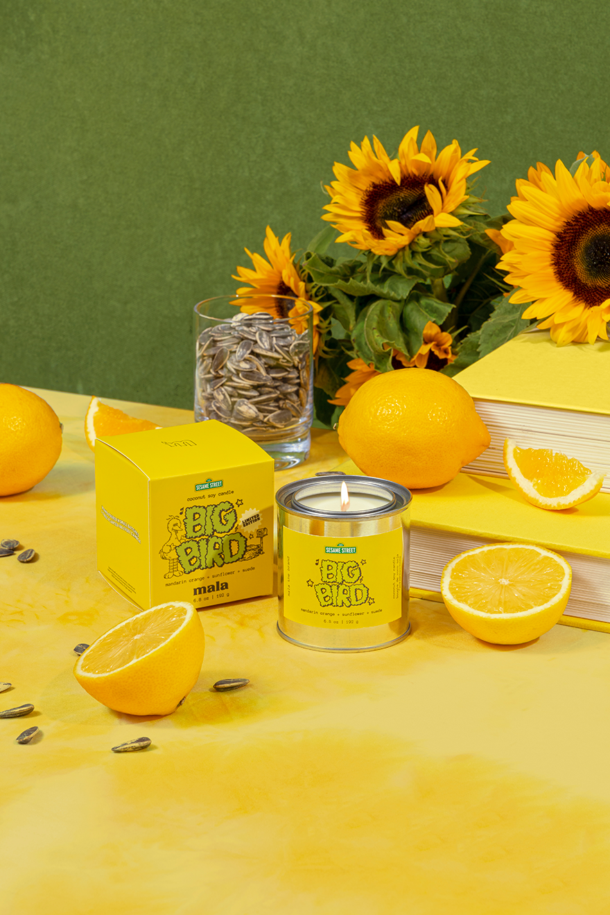 Yellow candle with sunflower seeds packaging on a table with sunflowers and oranges.