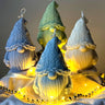 Four colorful Gnome-shaped candles on a wooden stand with string lights.