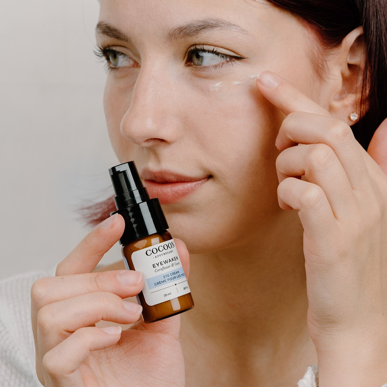 Woman applying eye cream with a bottle labeled 'COCOON' on a neutral background