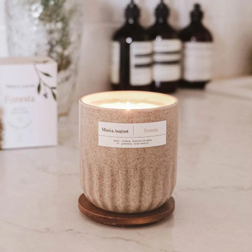 Candle in a textured container with a label on a marble surface, with bottles in the background. Foresta Reusable Candle by mimi and august
