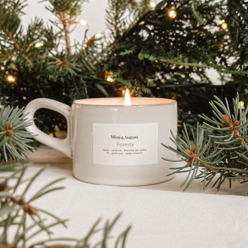 White mug candle with a label on a surface with Christmas tree branches and lights in the background Foresta Reusable Candle by mimi and august