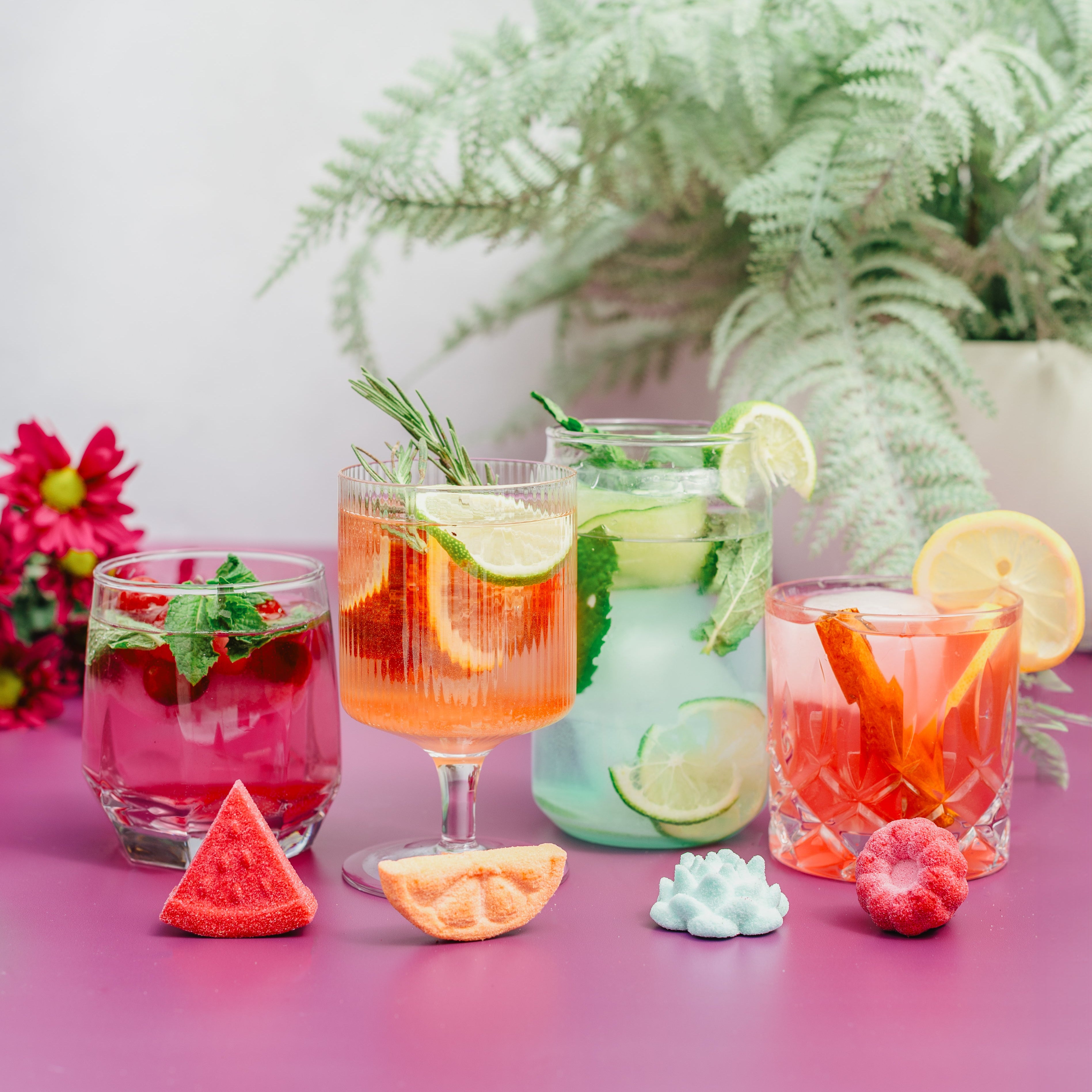 Assorted colorful cocktails with fruits and herbs on a pink surface with a plant in the background.