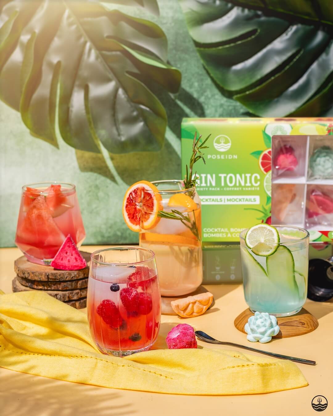 Assorted cocktails with garnishes and a box of tonics on a yellow cloth with green leafy background