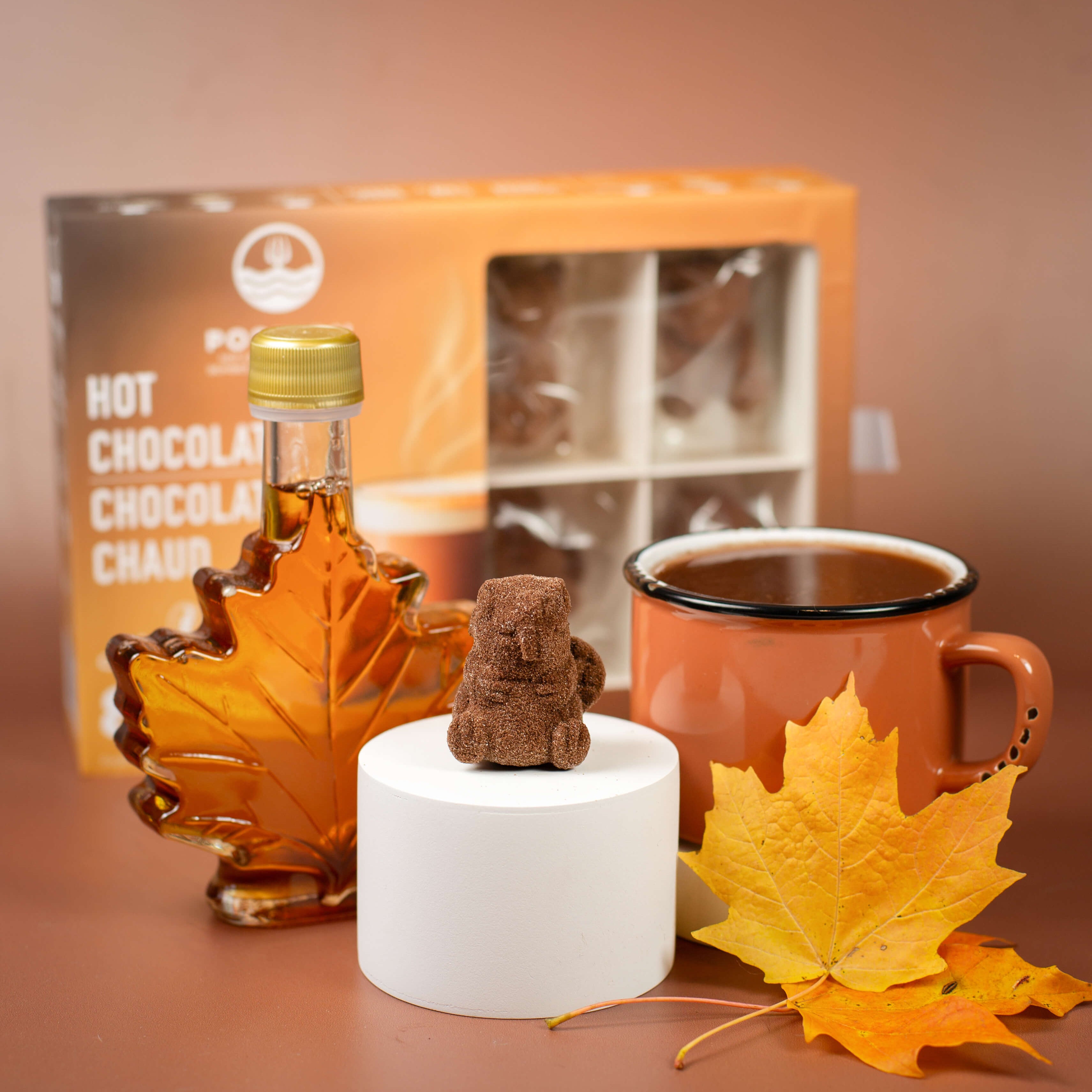 Maple syrup bottle, mug of hot chocolate, and decorative bear on a brown surface with a blurred background.