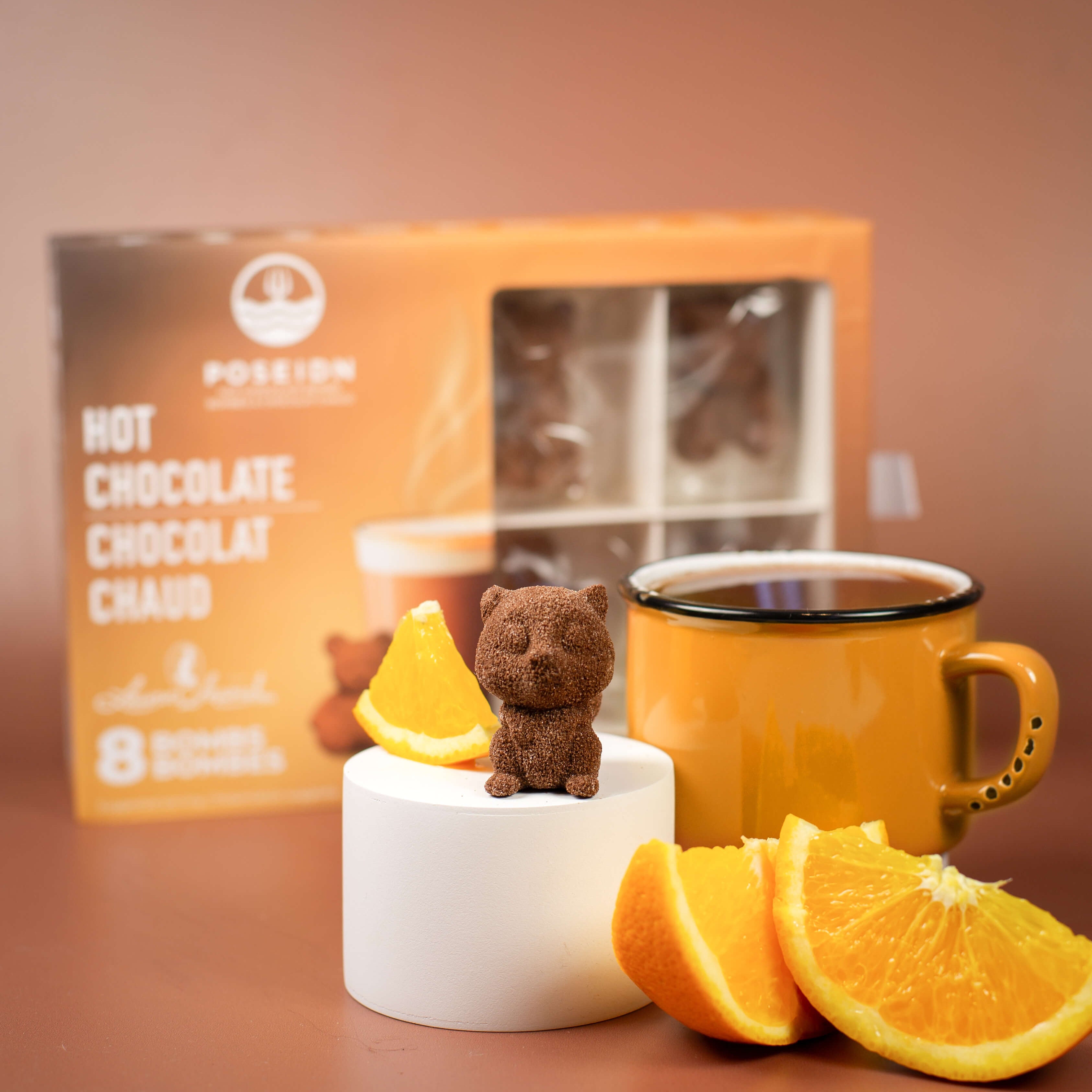Orange mug with a small bear figurine, orange slices, and a box of hot chocolate on a brown surface.