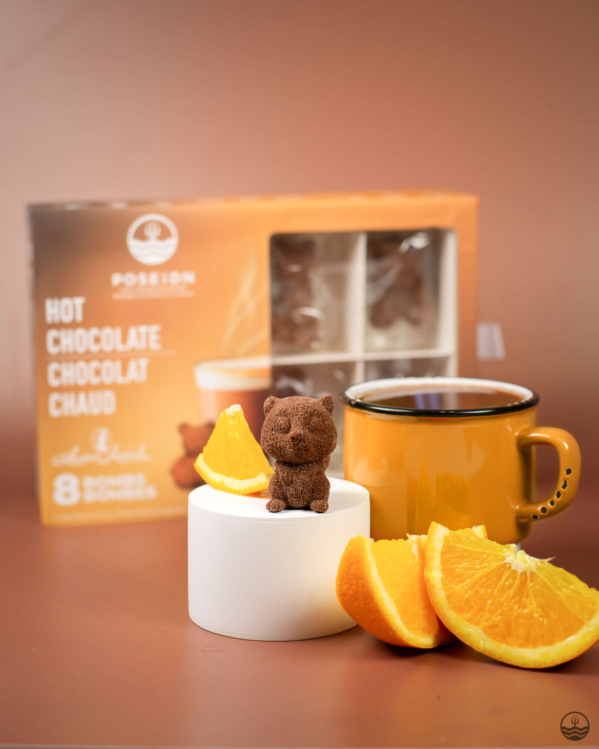 Orange mug with a small bear figurine, orange slices, and a box of hot chocolate on a brown surface.