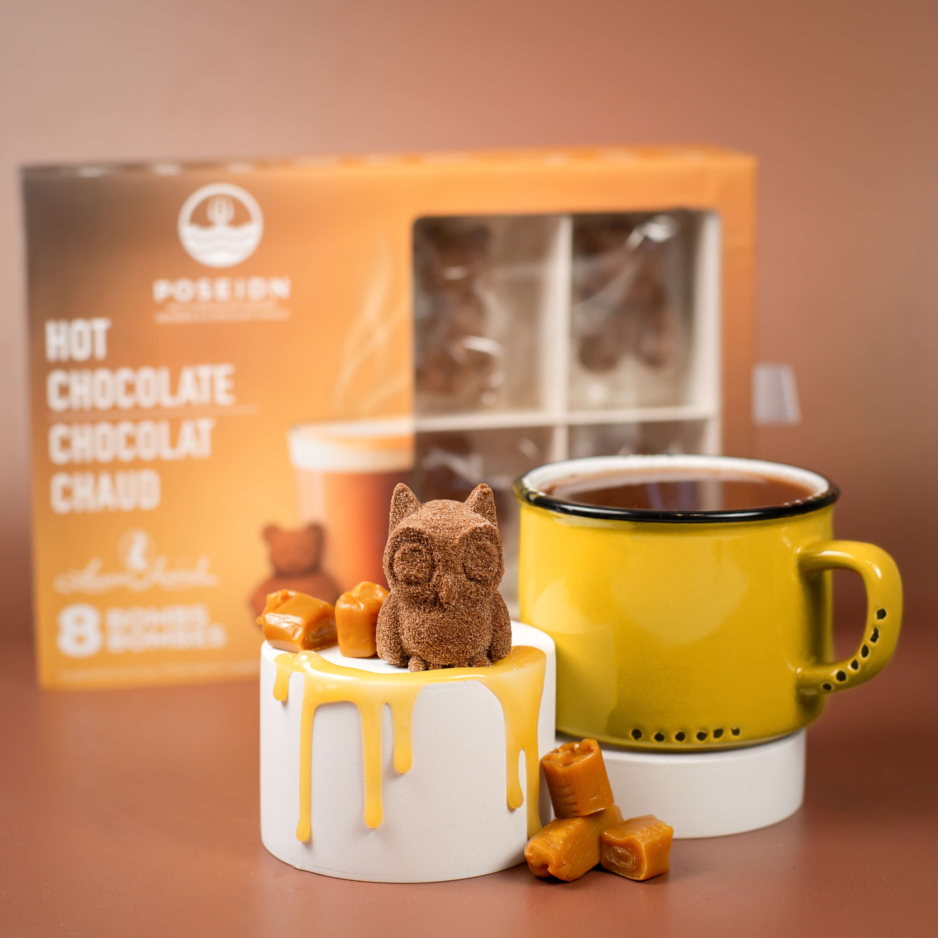 Yellow mug with a small owl-shaped chocolate bar and caramel pieces next to a box of hot chocolate.