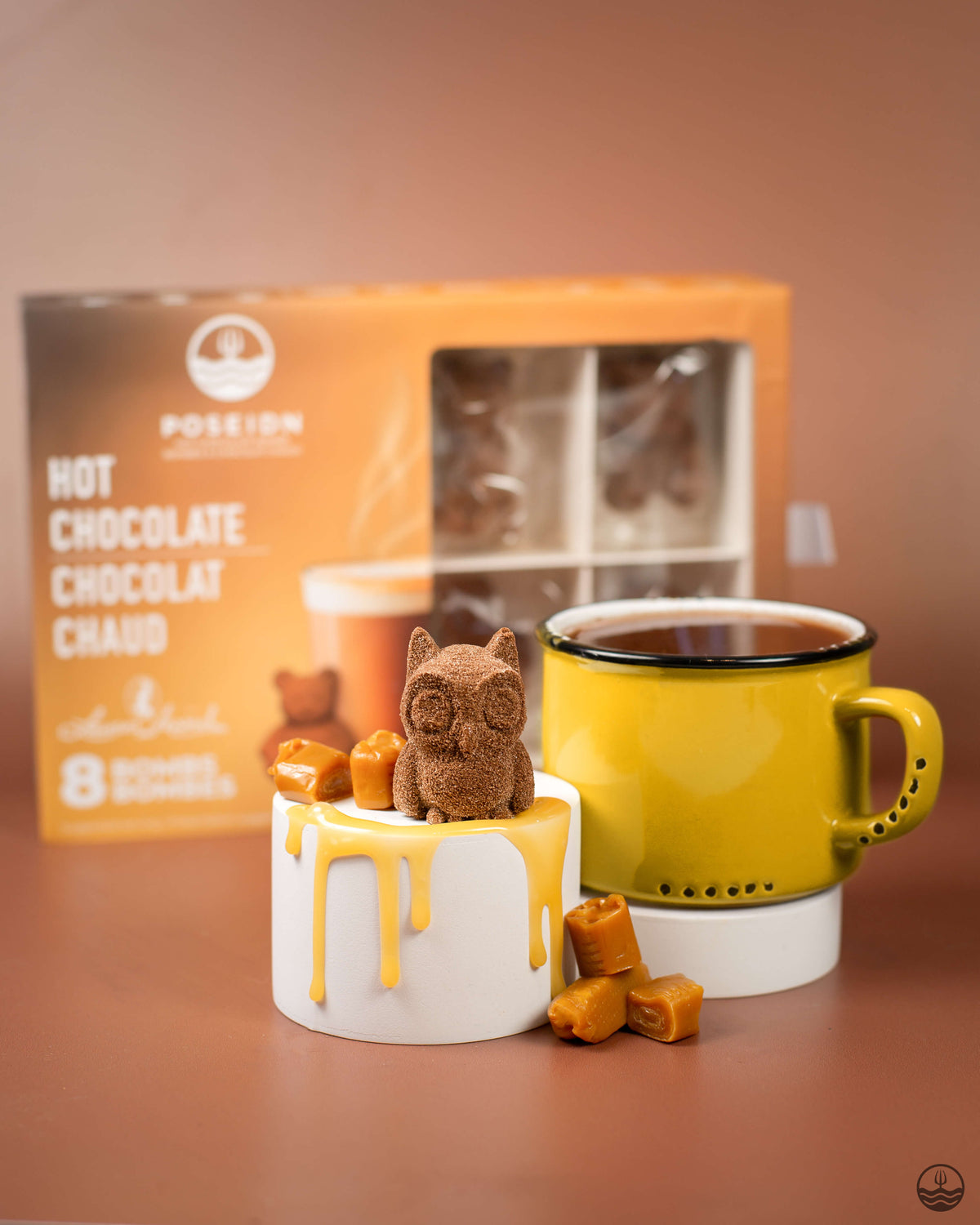 Yellow mug with a small owl-shaped chocolate bar and caramel pieces next to a box of hot chocolate.